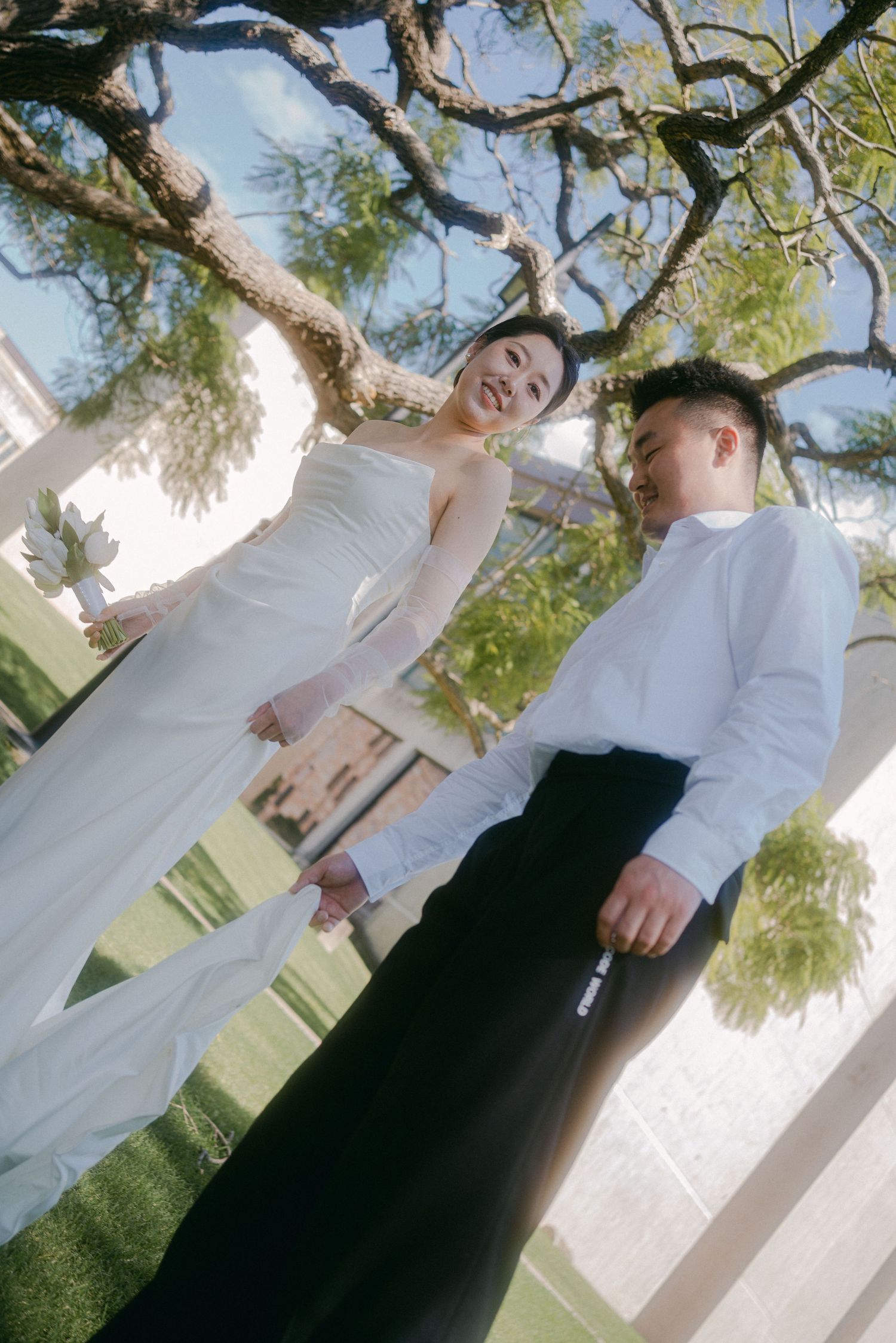A couple in formal wedding attire poses outdoors under a tree with white columns visible in the background.