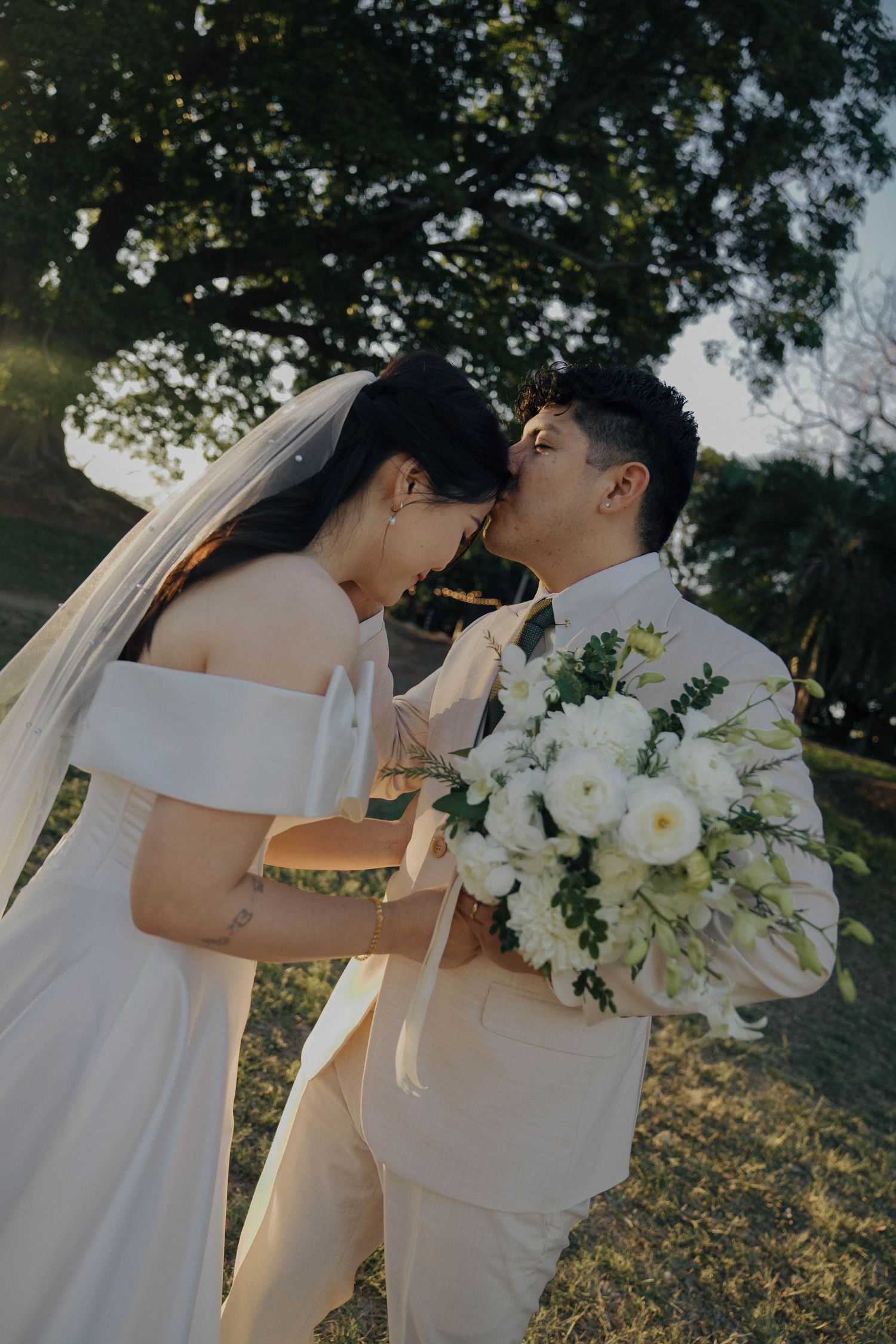 A romantic wedding couple shares intimate moments in an outdoor garden setting with white floral bouquet and elegant attire.