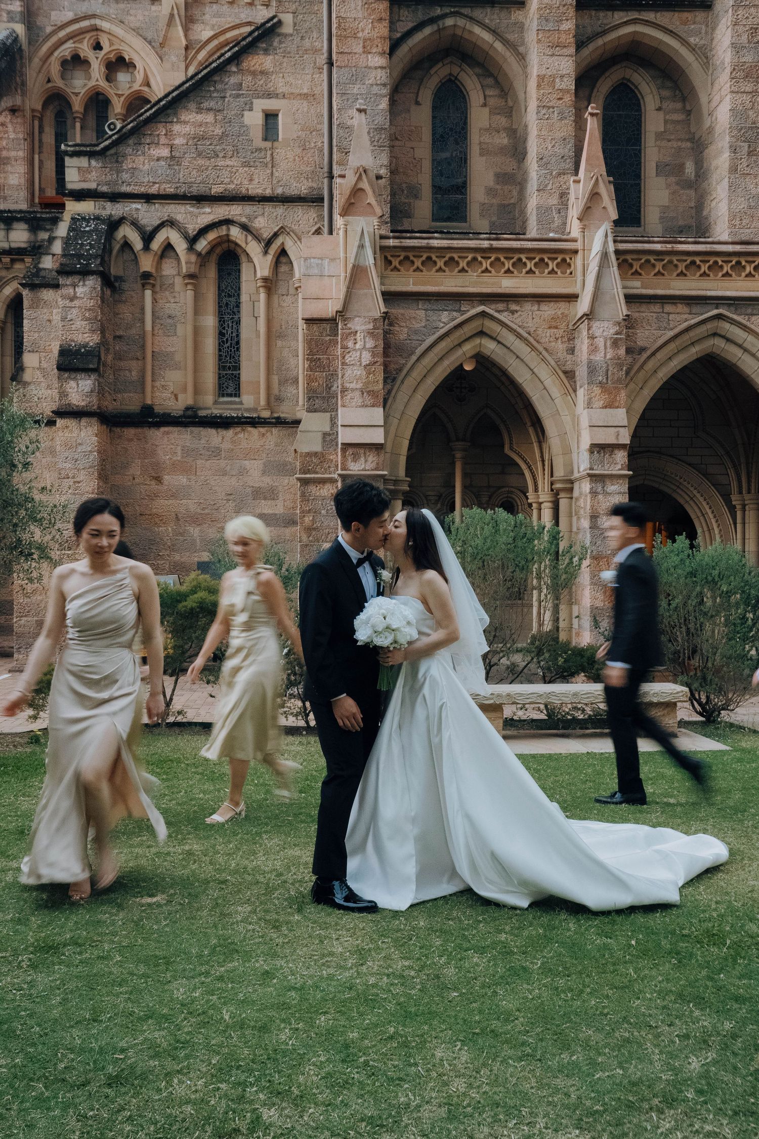 Wedding party members playfully fall onto grass in sequence outside a gothic stone church building.