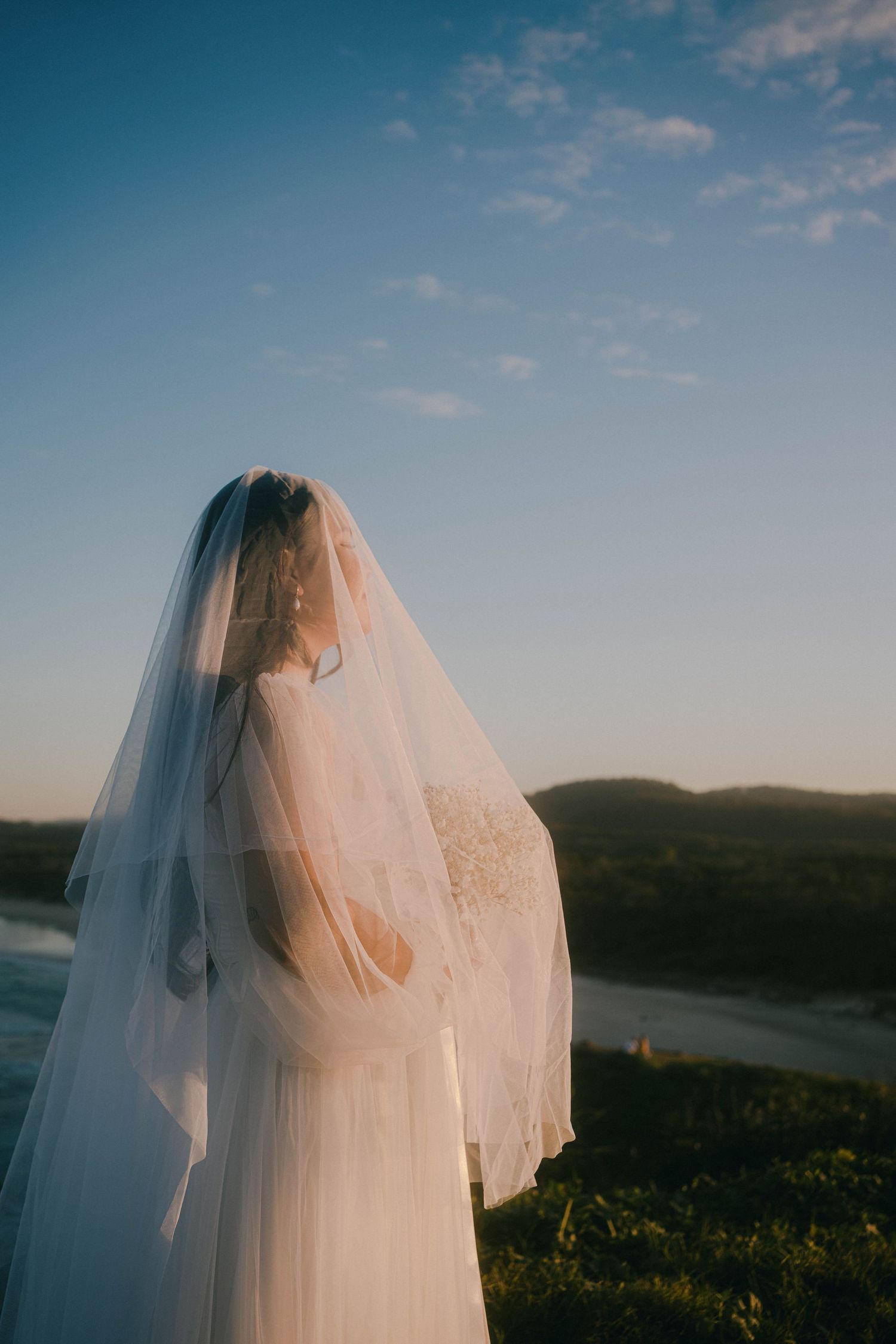 A bride in a flowing white veil stands silhouetted against a scenic mountain landscape at sunset.