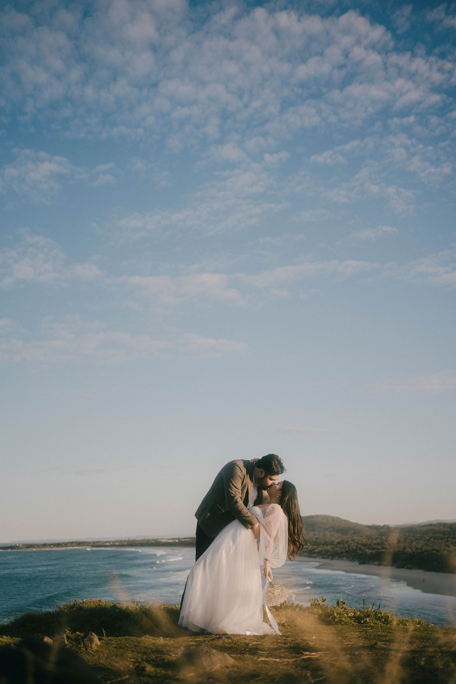 A couple in wedding attire embraces on a scenic coastal cliff overlooking the ocean under a bright blue sky.