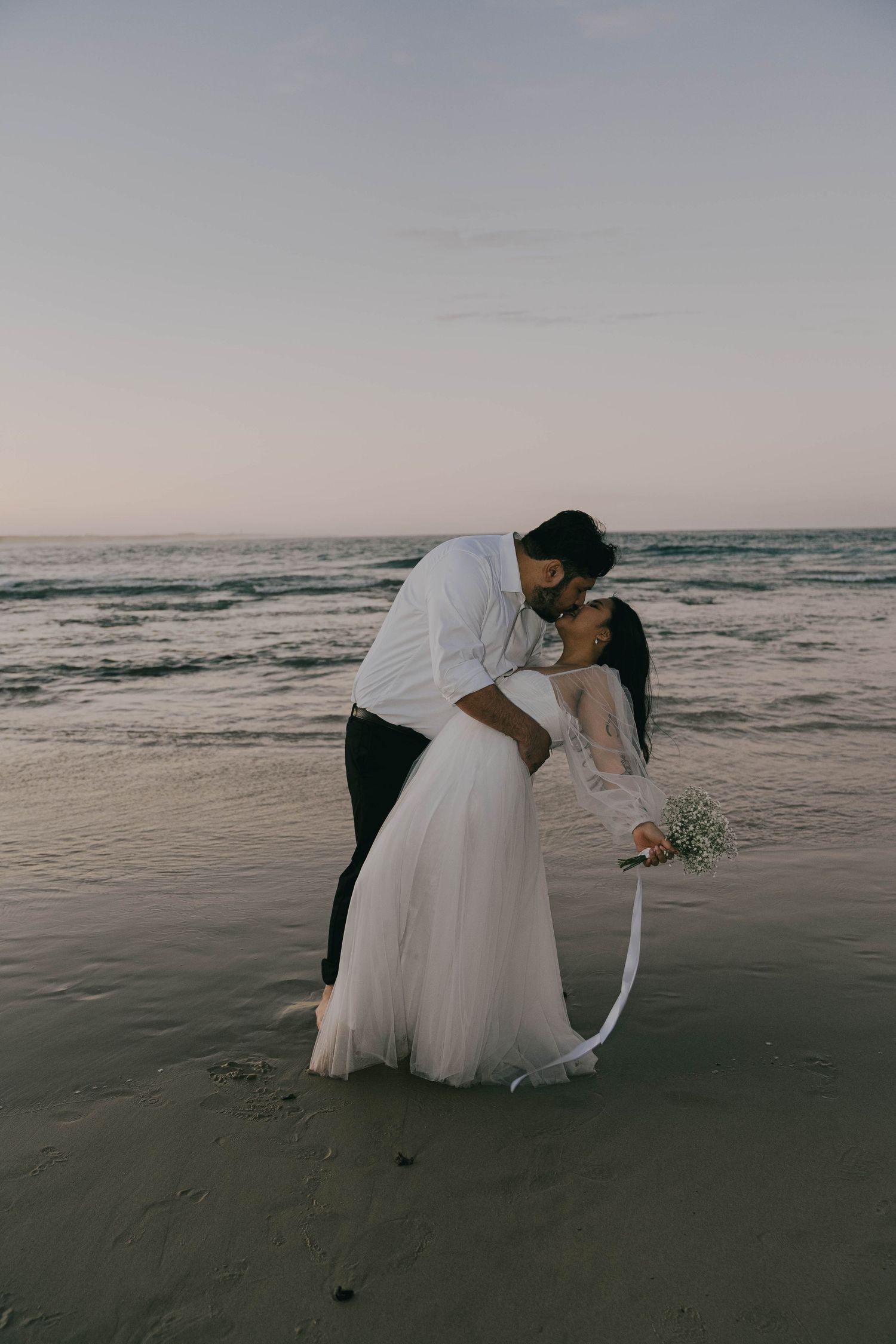 A couple in wedding attire shares a romantic moment on a moody beach at dusk with waves in the background.