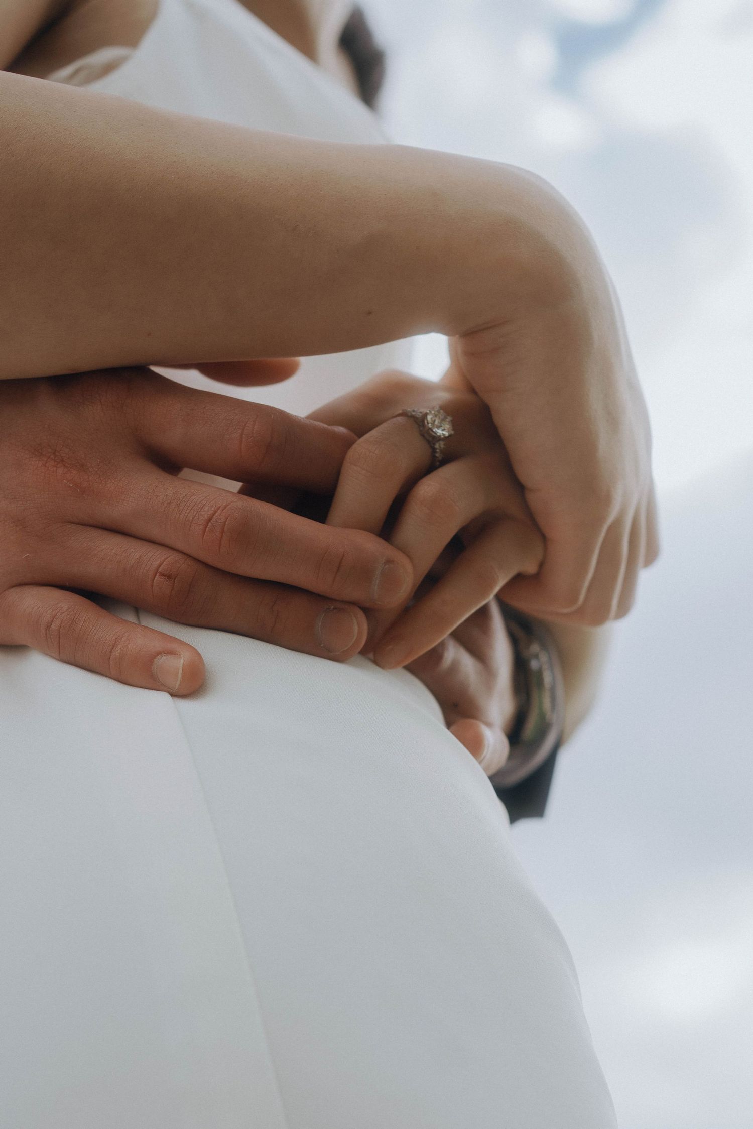 Close-up shot of hands with engagement ring against white fabric and soft cloudy background.