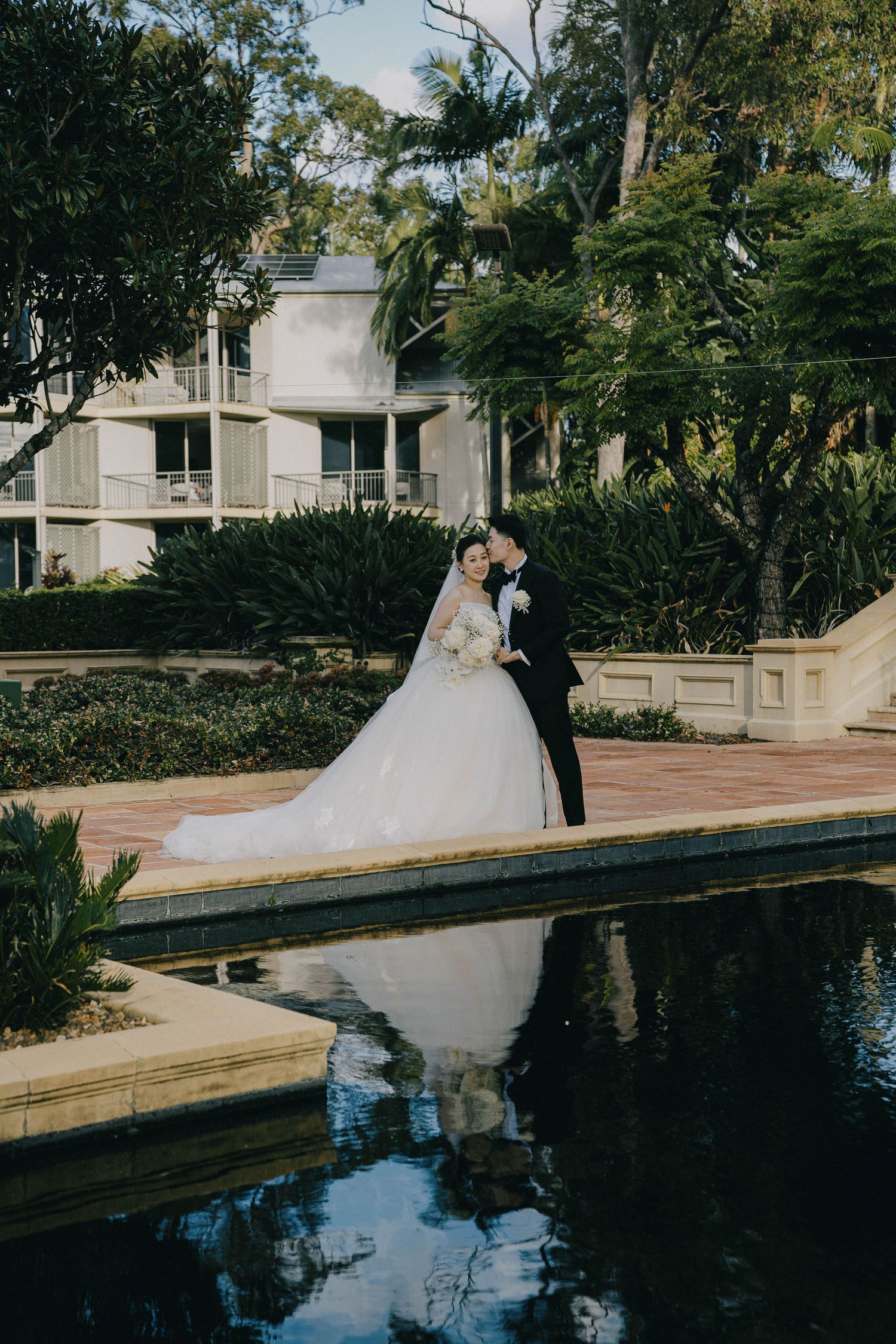 Elegant couple in wedding attire pose beside a reflective pool surrounded by lush gardens at a luxurious venue.