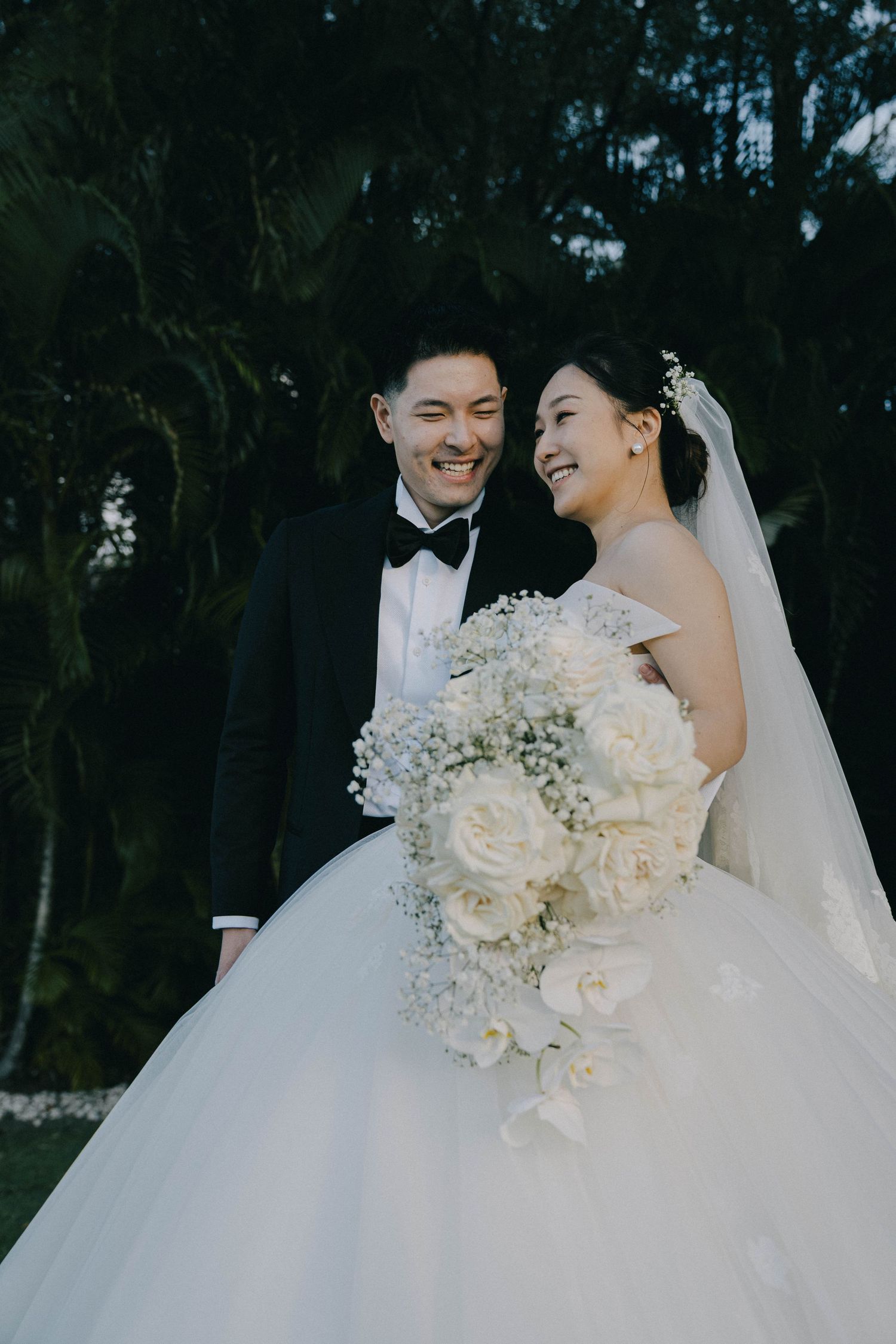A joyful wedding couple shares a moment outdoors, with the bride holding an elegant white and baby's breath bouquet.