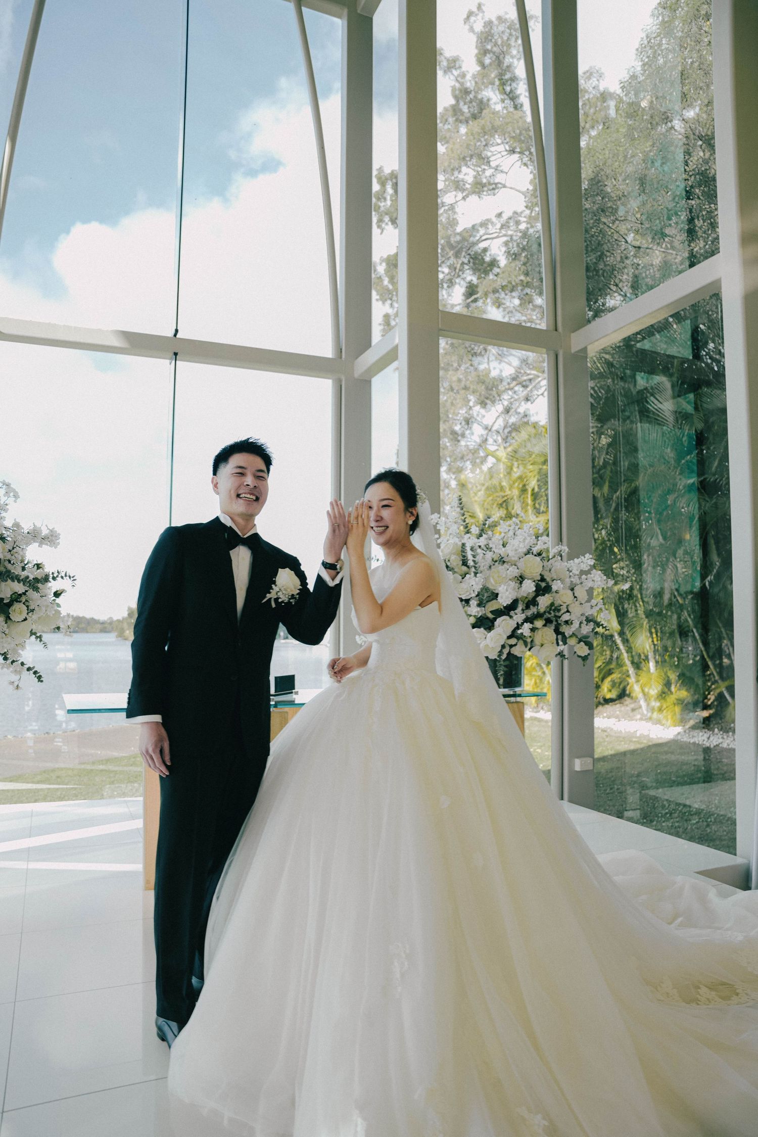 A bride and groom share intimate moments in a bright glass-walled venue with natural light streaming through the windows.