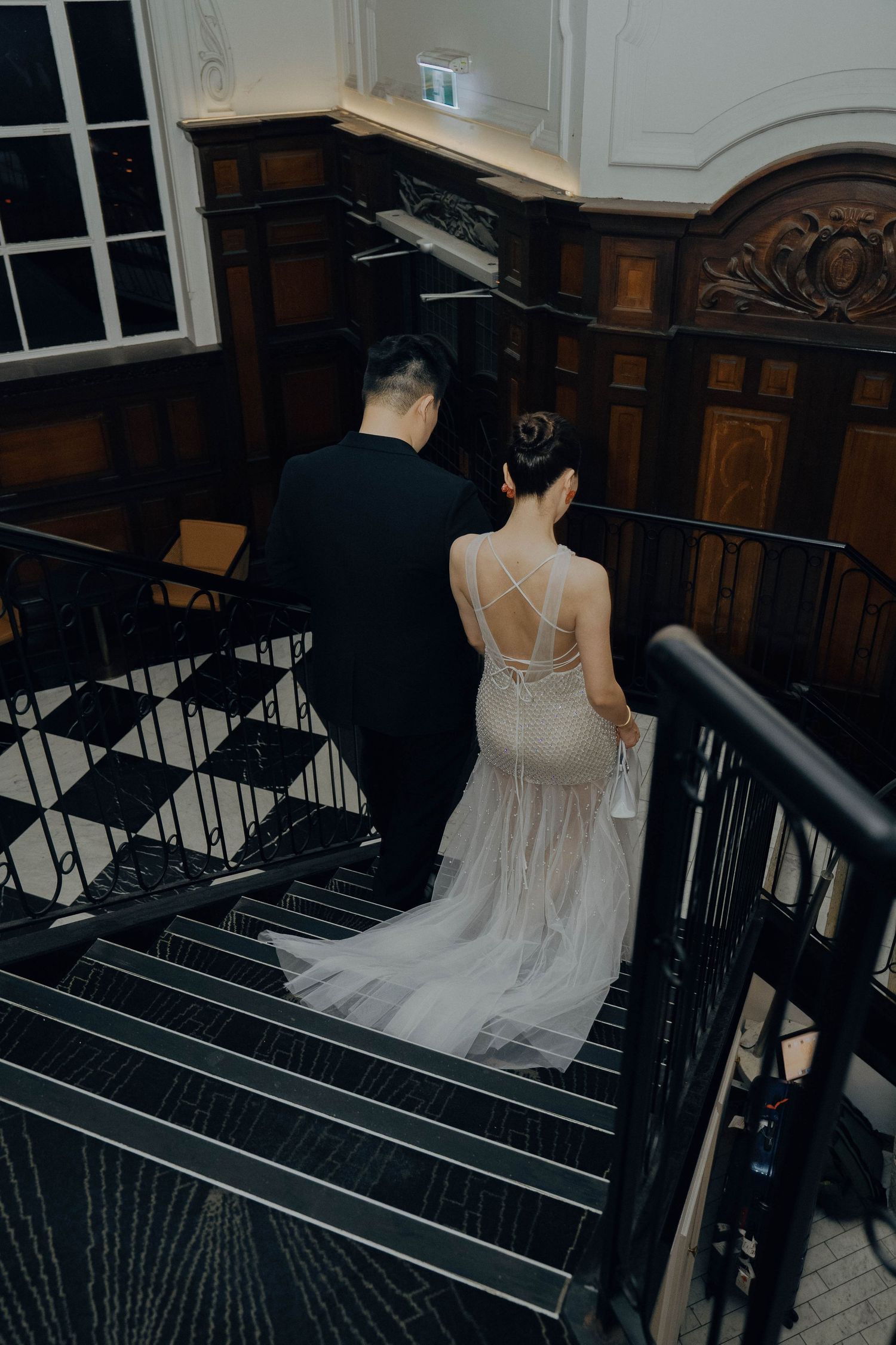 A couple descending an elegant staircase, with the bride wearing a sheer white backless gown with delicate straps.