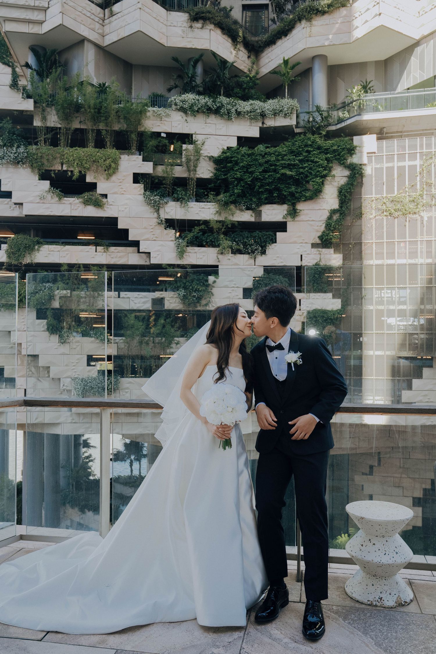 A bride and groom share an intimate moment in front of a modern architectural backdrop with cascading greenery.
