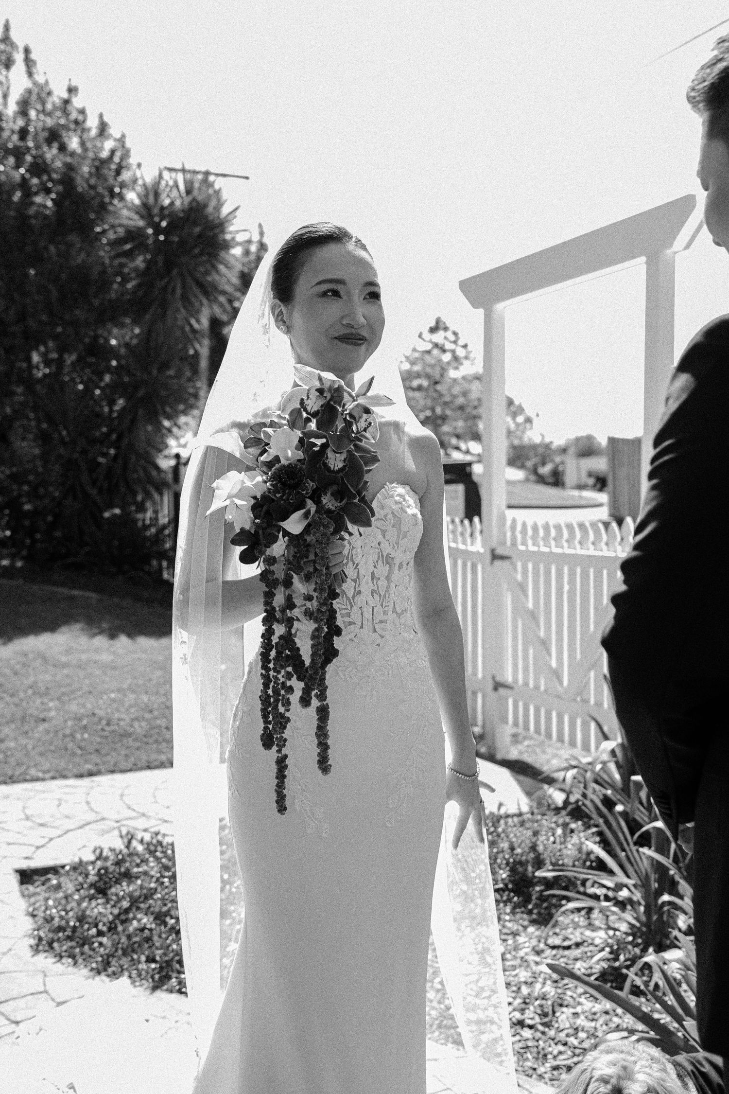 Black and white wedding photo showing bride in elegant fitted lace gown with veil holding cascading floral bouquet on porch.