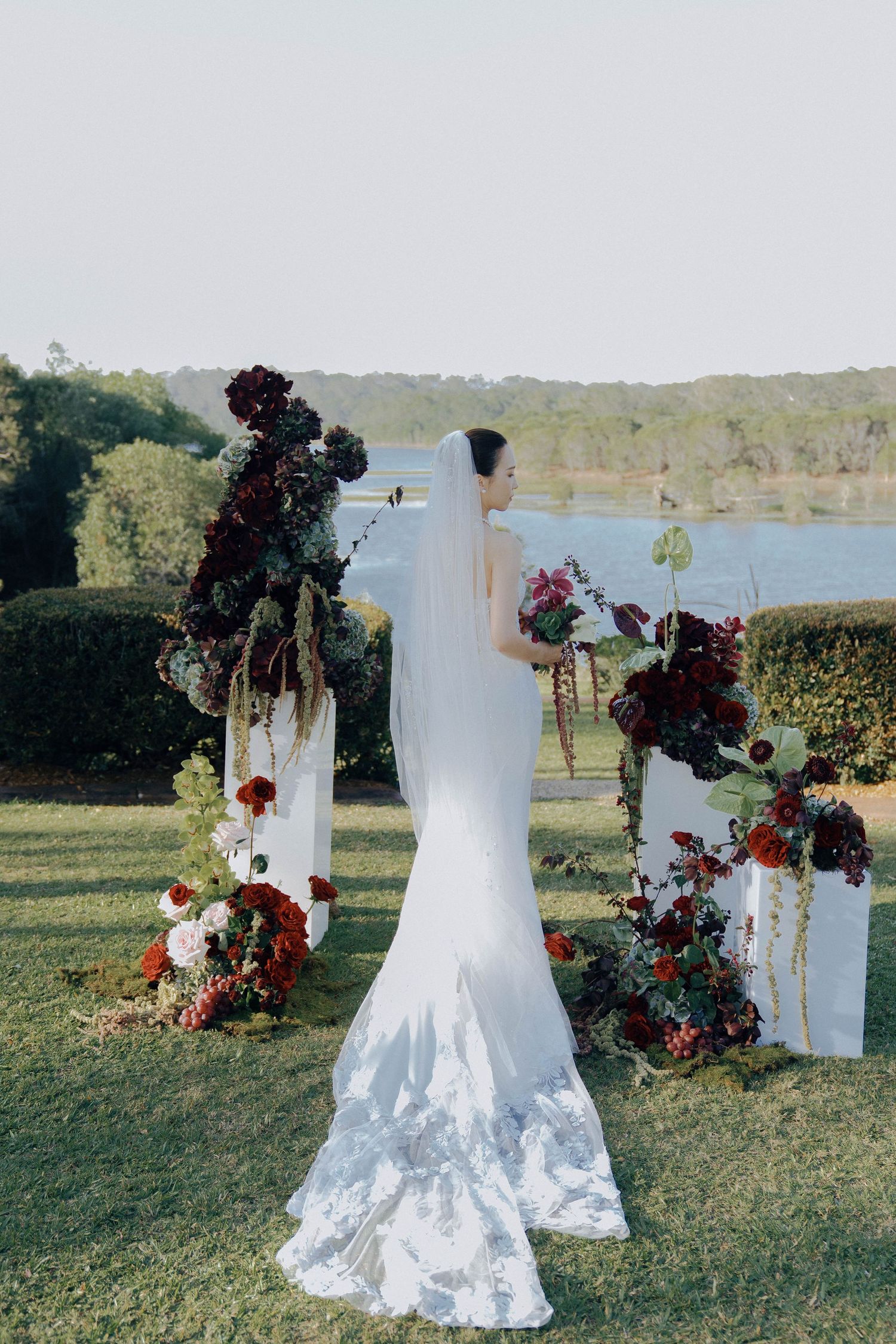 White wedding dress on display outdoors with deep burgundy floral arrangements against a scenic lake backdrop.