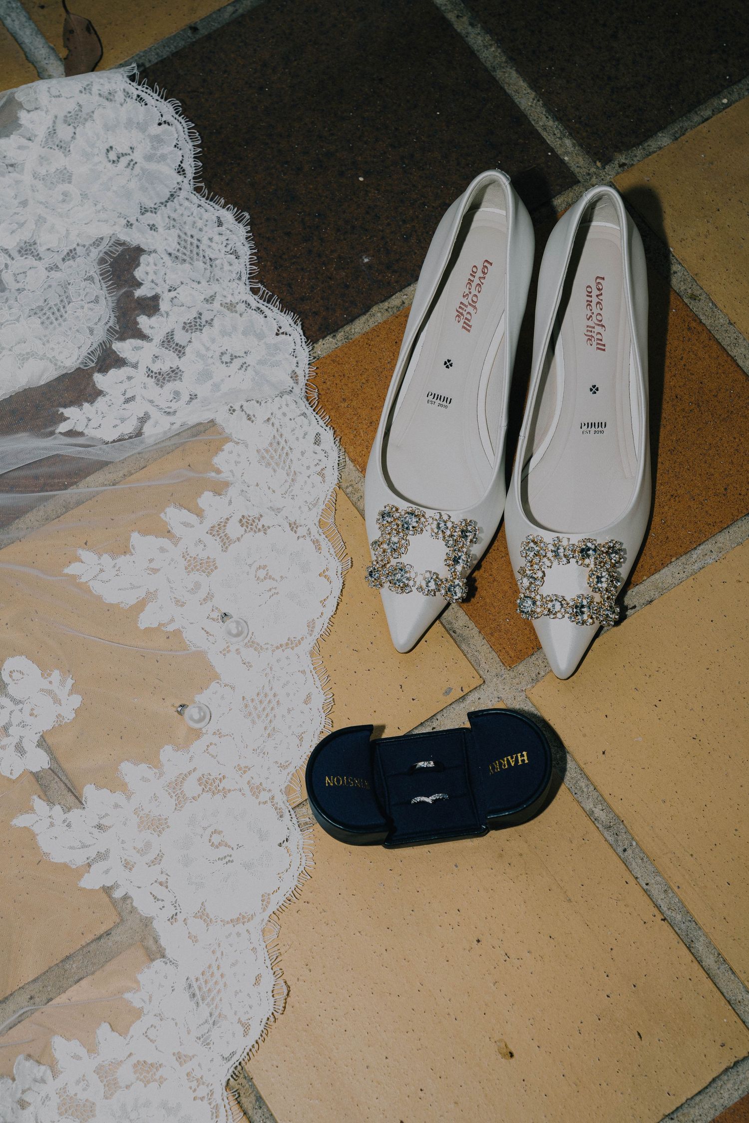 White lace fabric lies on a beige tile floor next to white pointed-toe shoes with decorative details.