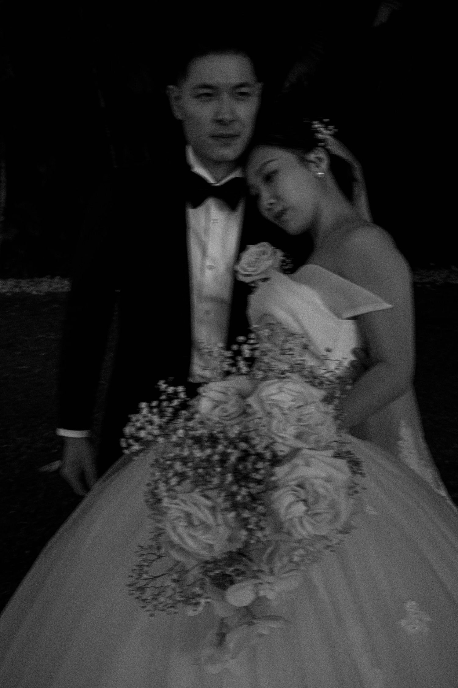 A couple in formal wedding attire stands together while holding an elegant white floral bouquet.