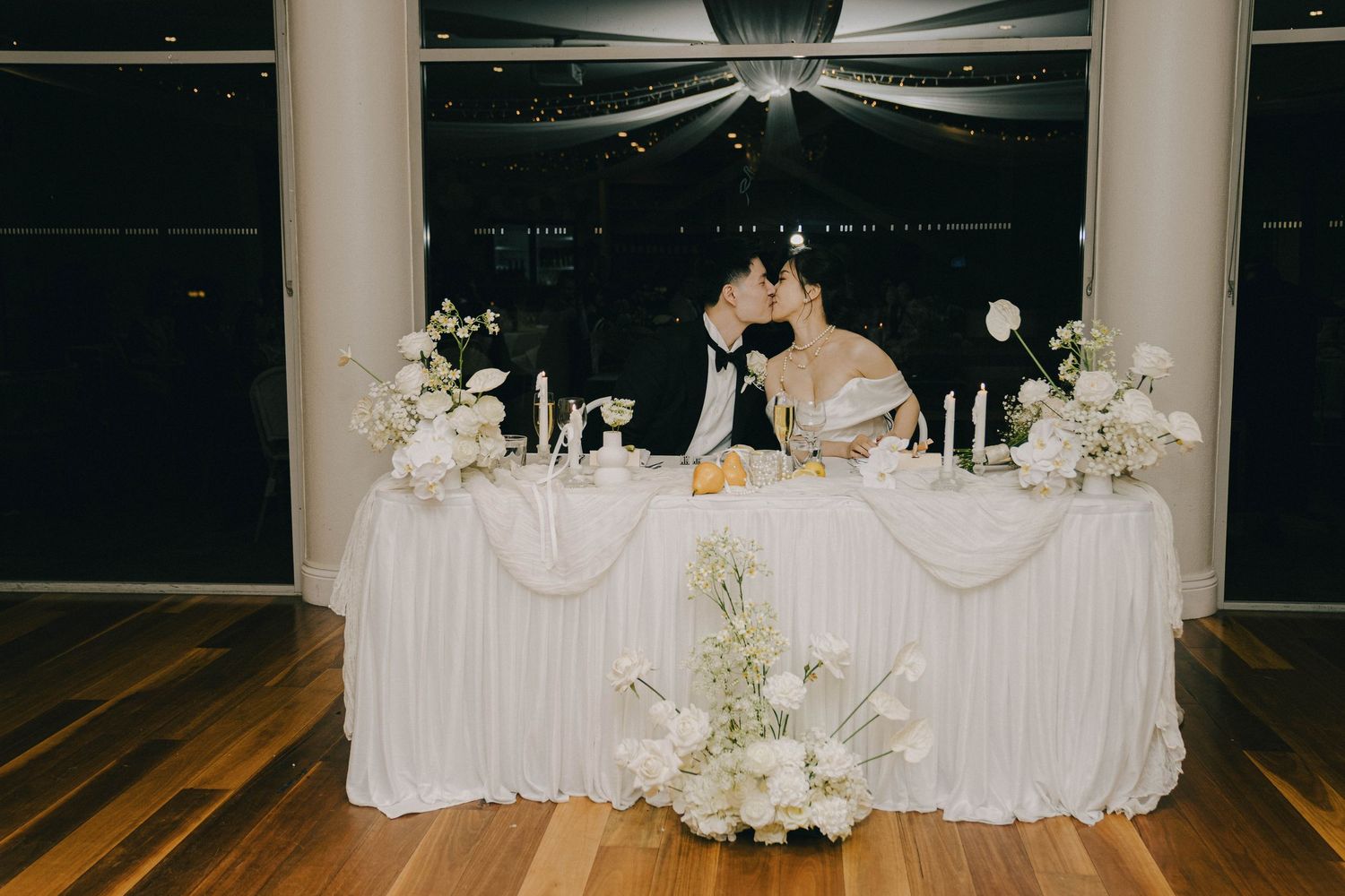 Romantic couple shares a kiss at elegant wedding reception head table decorated with white flowers and drapery.