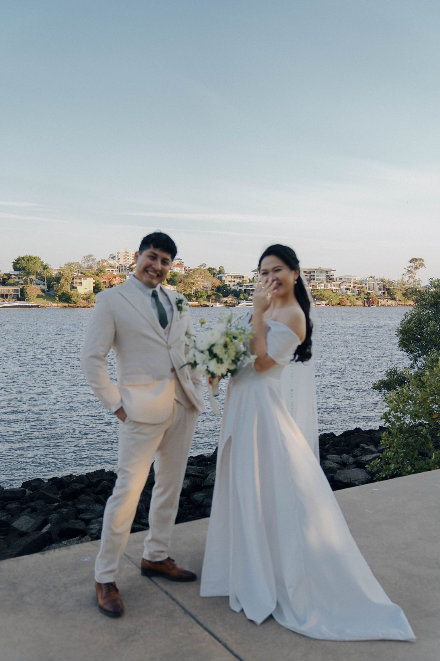 A couple in wedding attire poses together on a rocky beach at sunset with water and trees in the background.