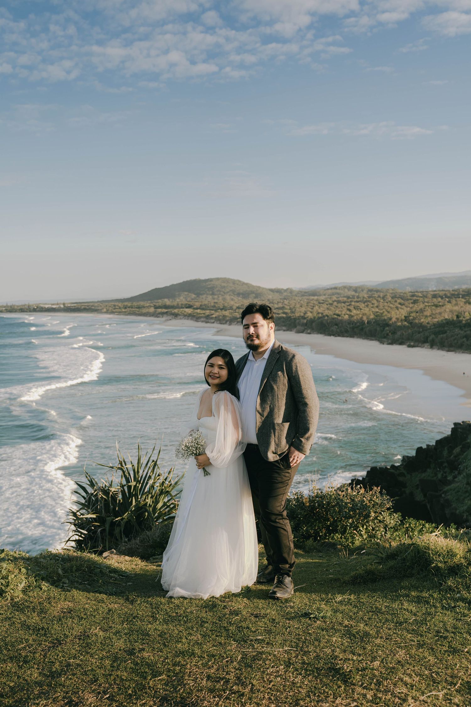 Scenic coastal wedding photos taken at a hilltop overlooking a pristine beach with rolling waves and mountains in the distance.