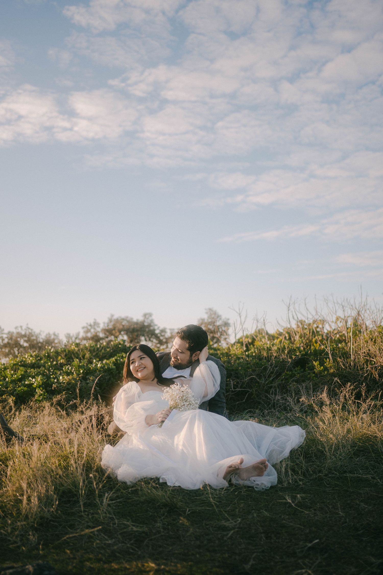 A couple in wedding attire sits together on a grassy hillside during golden hour with wispy clouds in the sky.