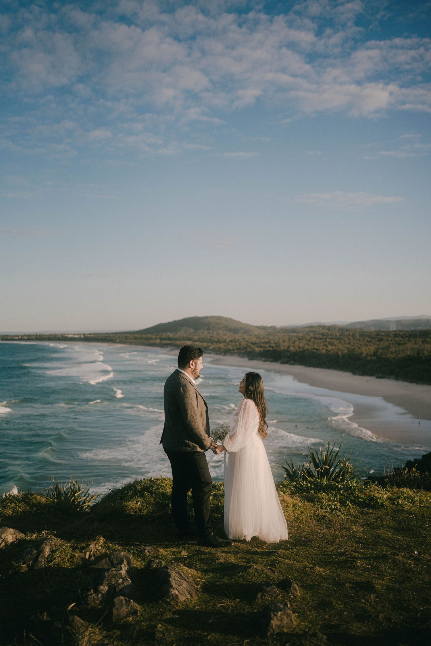 A couple stands on a coastal cliff overlooking a scenic beach with rolling waves and mountains in the distance.