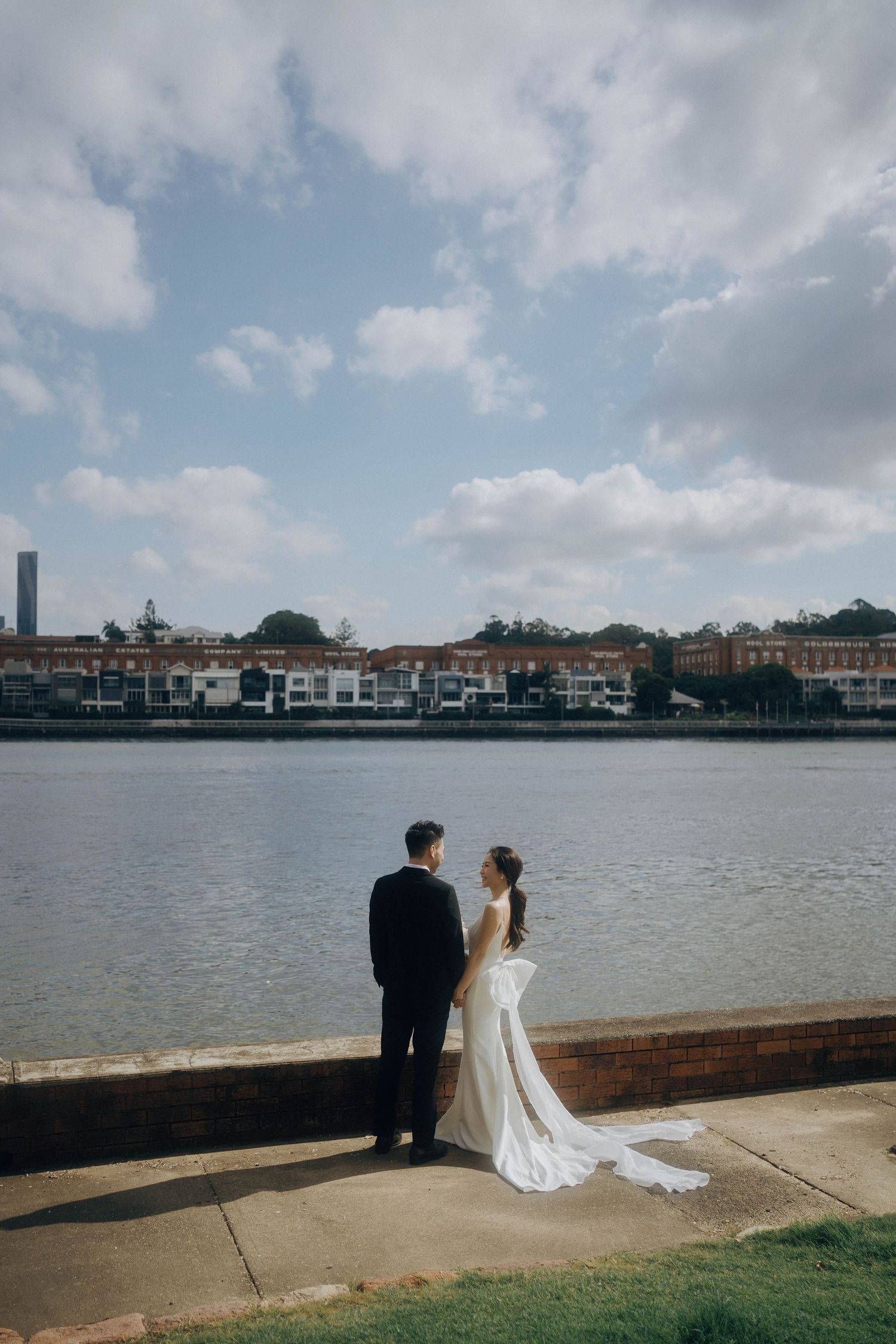 A couple in wedding attire sits by a waterfront with buildings and cloudy skies in the background.