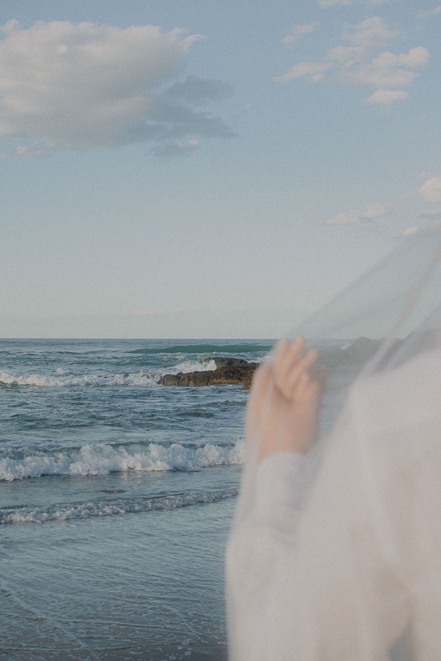 A person in white clothing stands by the ocean shore as gentle waves roll in under a serene blue sky.