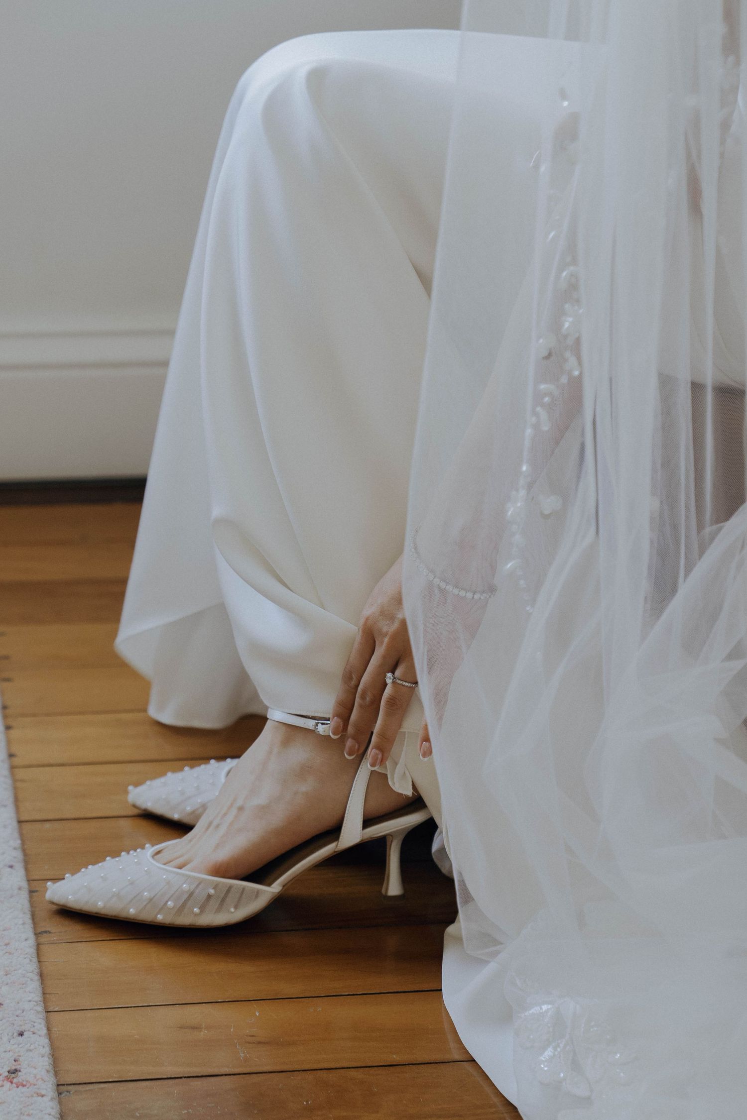 White bridal shoes with delicate beading peek out from under a flowing wedding dress and veil on hardwood flooring.