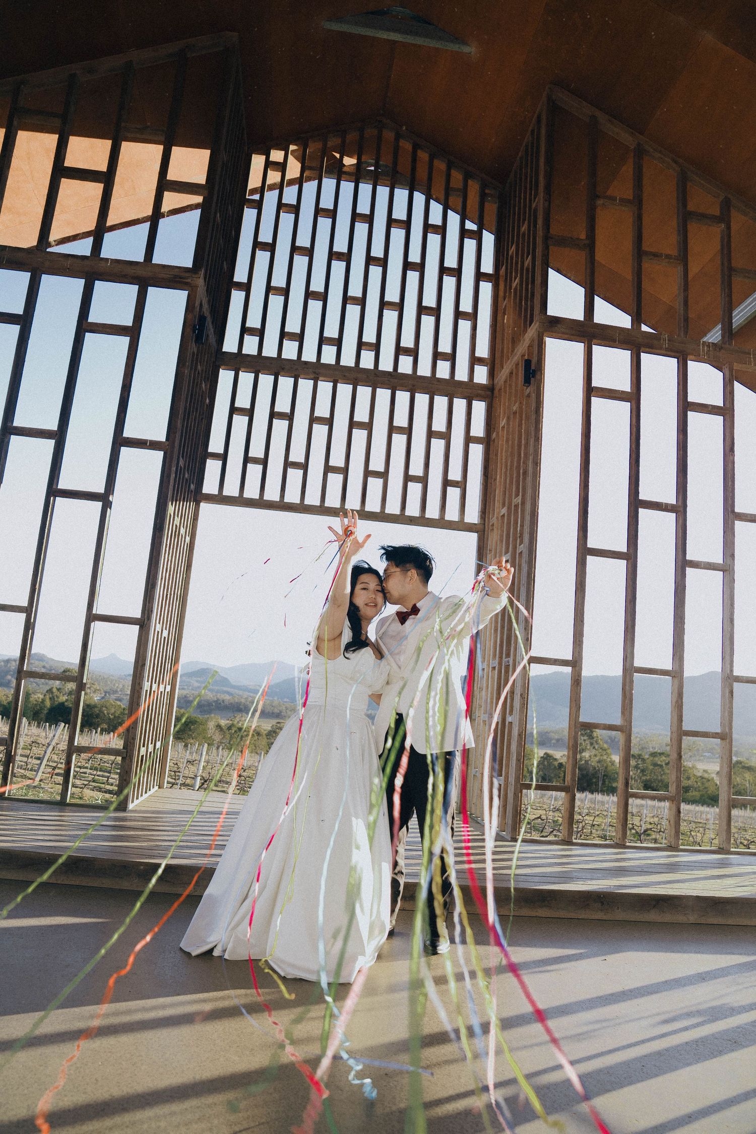Couple celebrates in elegant wedding attire at a chapel with mountain views through geometric windows and colorful ribbons.