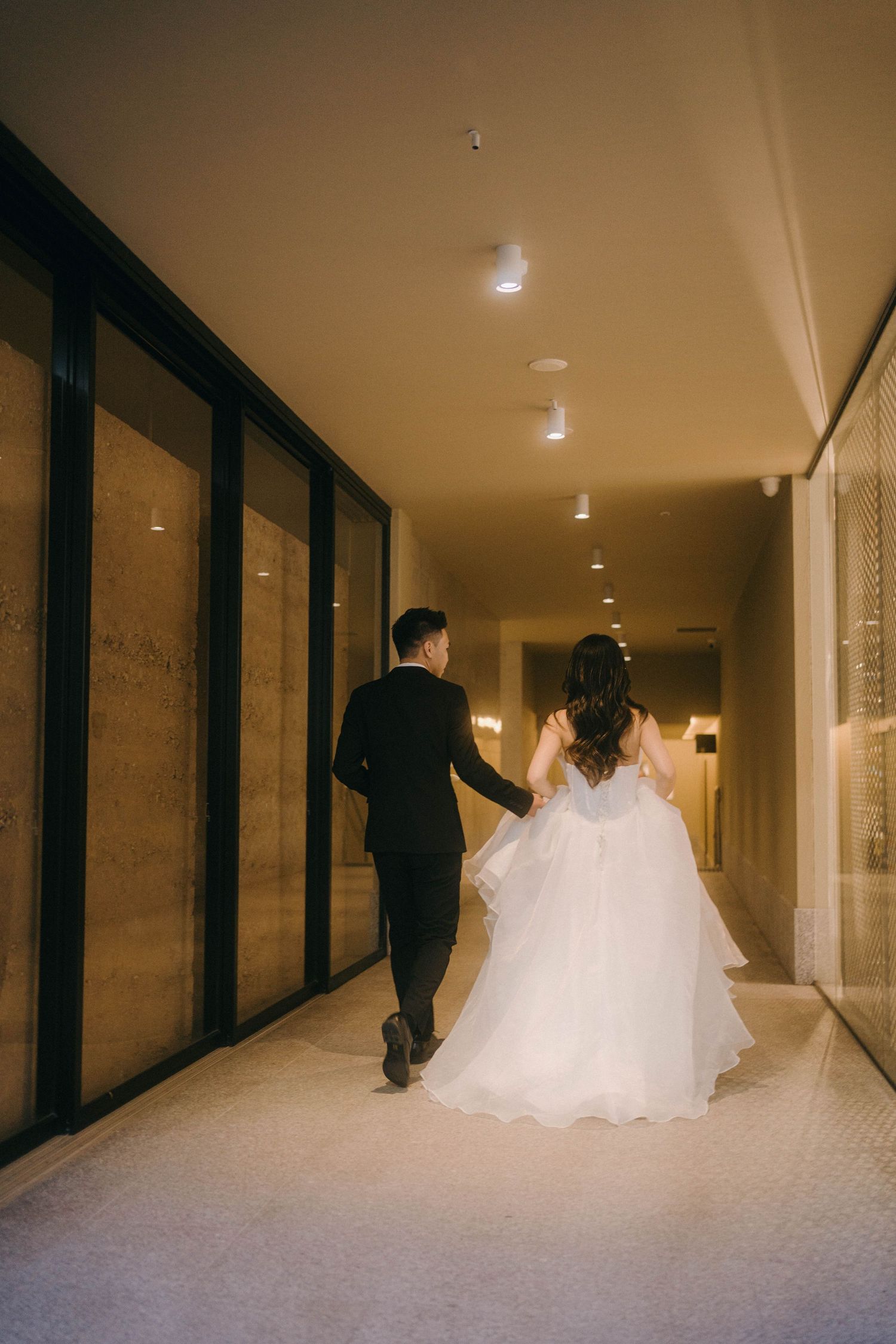 A couple in wedding attire walks down an elegant hallway with warm lighting and glass walls.