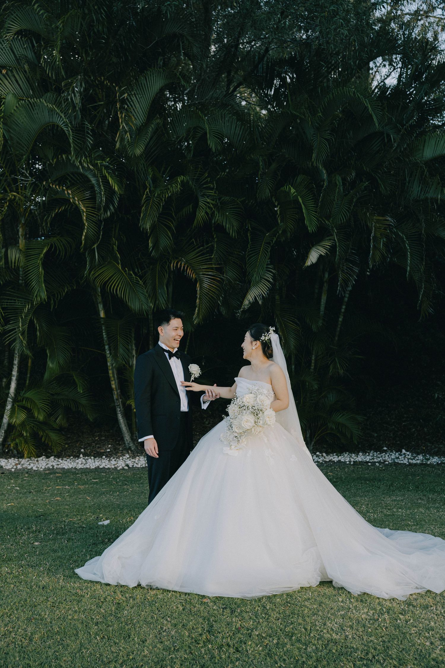 A wedding couple poses in front of lush tropical palm trees on a manicured lawn during their outdoor ceremony.