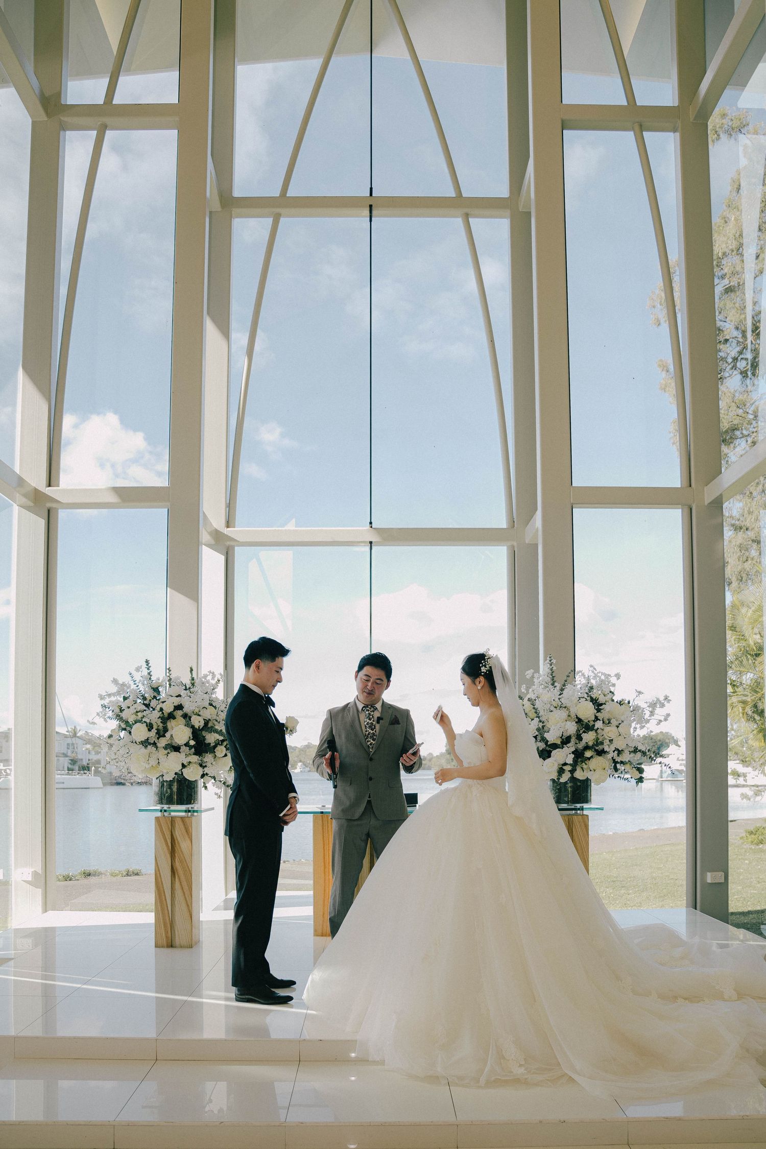 A wedding ceremony takes place in a bright, modern chapel with floor-to-ceiling windows and white decor.
