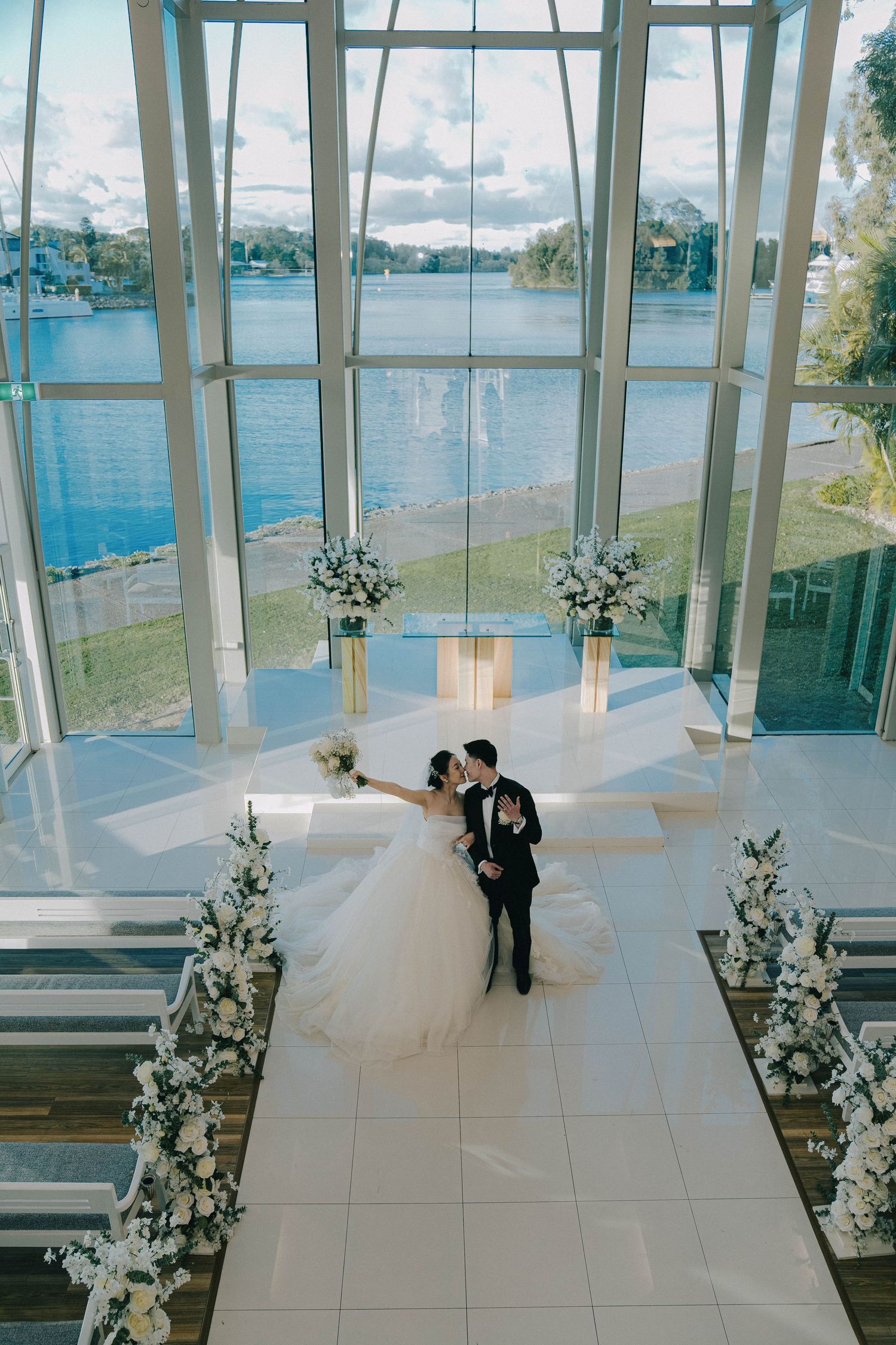 A couple shares an intimate moment in a modern glass chapel with white floral arrangements and a lakeside view.