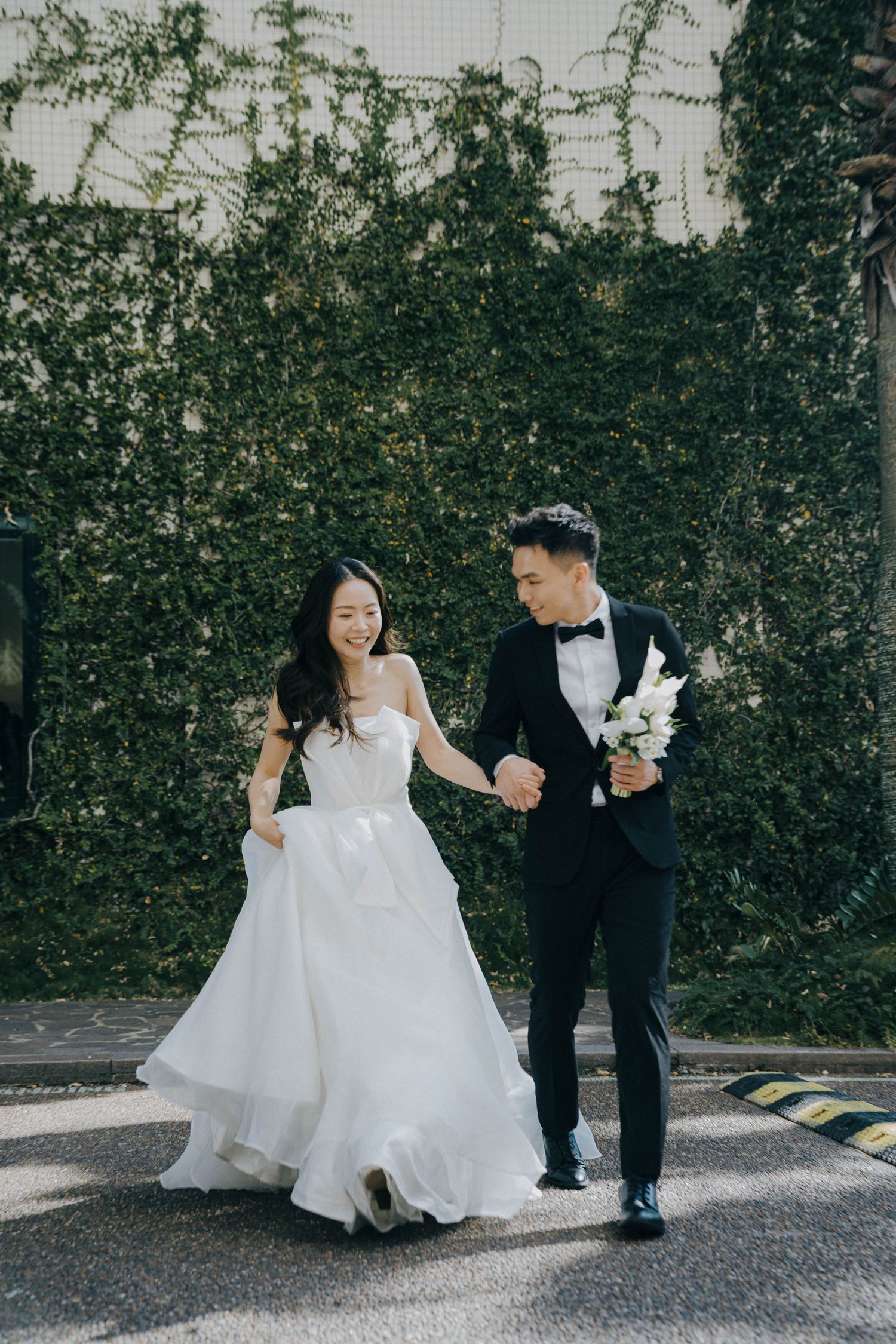 A couple in wedding attire shares playful moments while walking together along a path with greenery in the background.