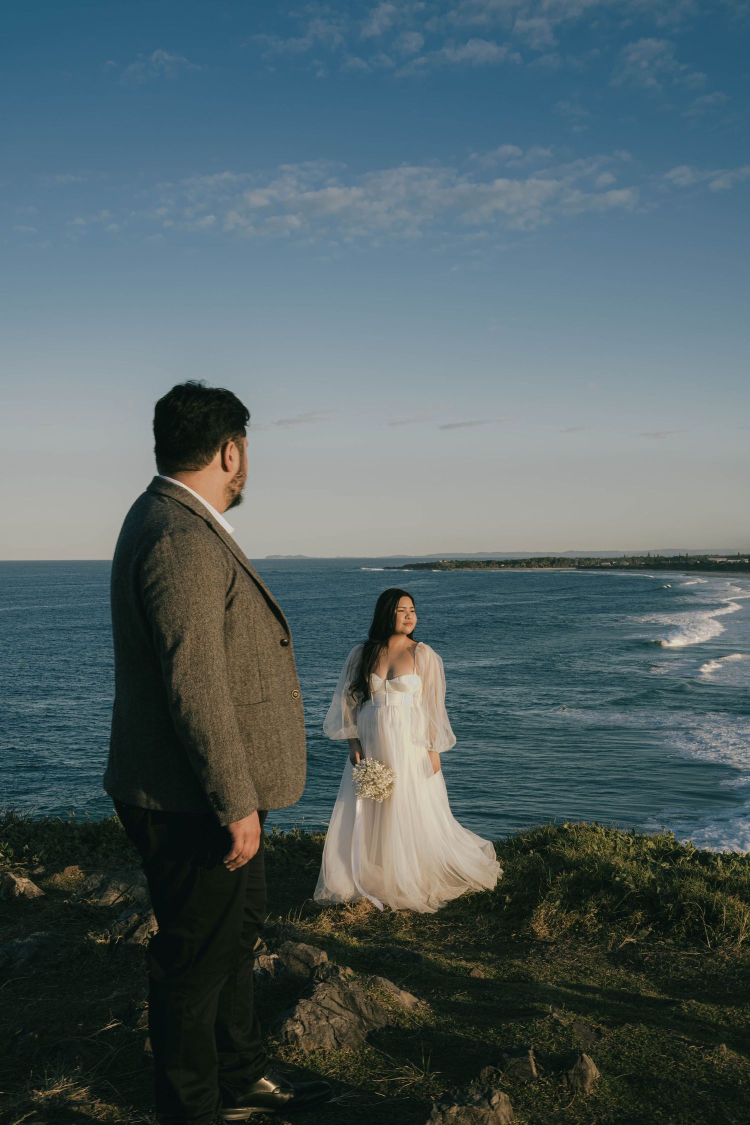 A couple in wedding attire stands on rocky coastal cliffs overlooking dramatic ocean waves and blue skies.