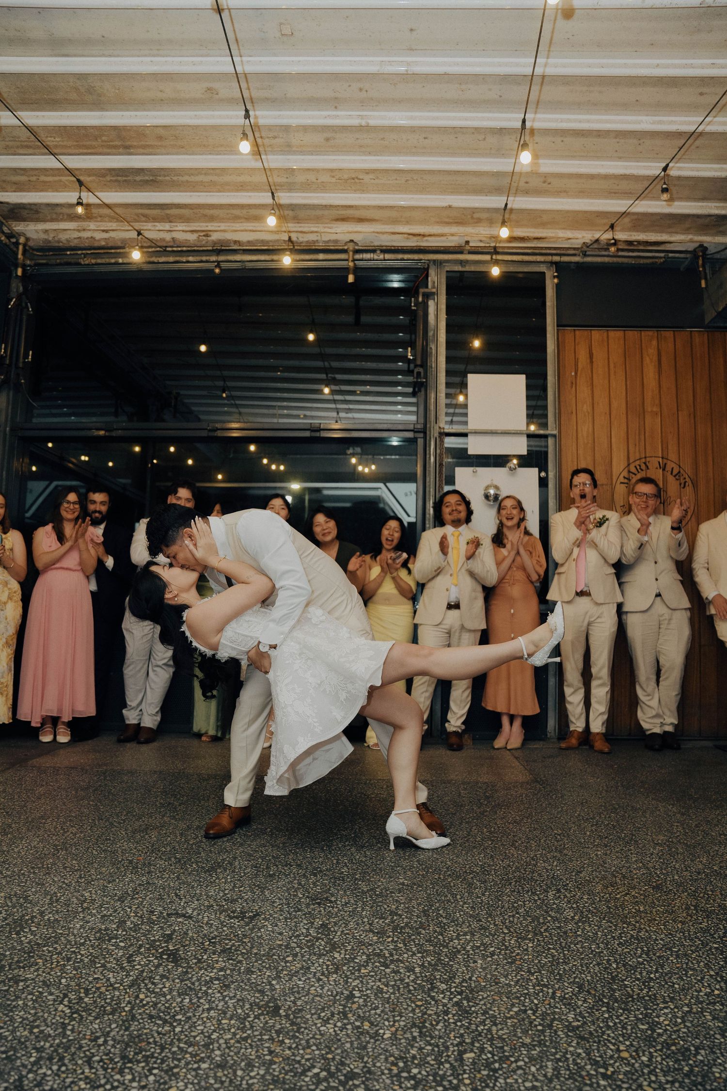 A couple performs a romantic dip dance move at their wedding reception while guests in pink dresses and beige suits watch.