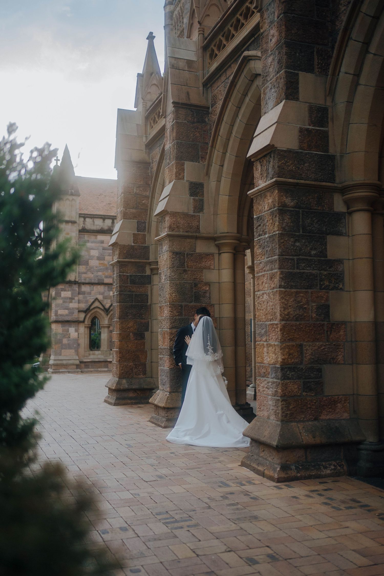 A wedding dress and veil silhouetted against gothic stone church archways and columns on a brick pathway.