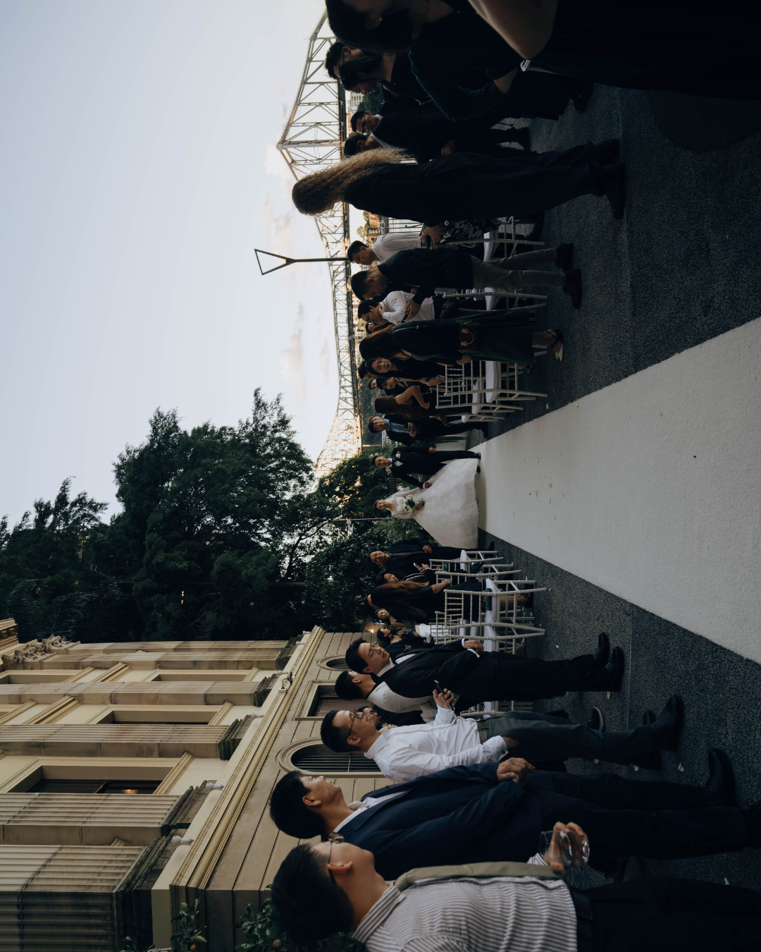 People lined up in dark clothing stand against a railing on what appears to be a train platform or walkway.