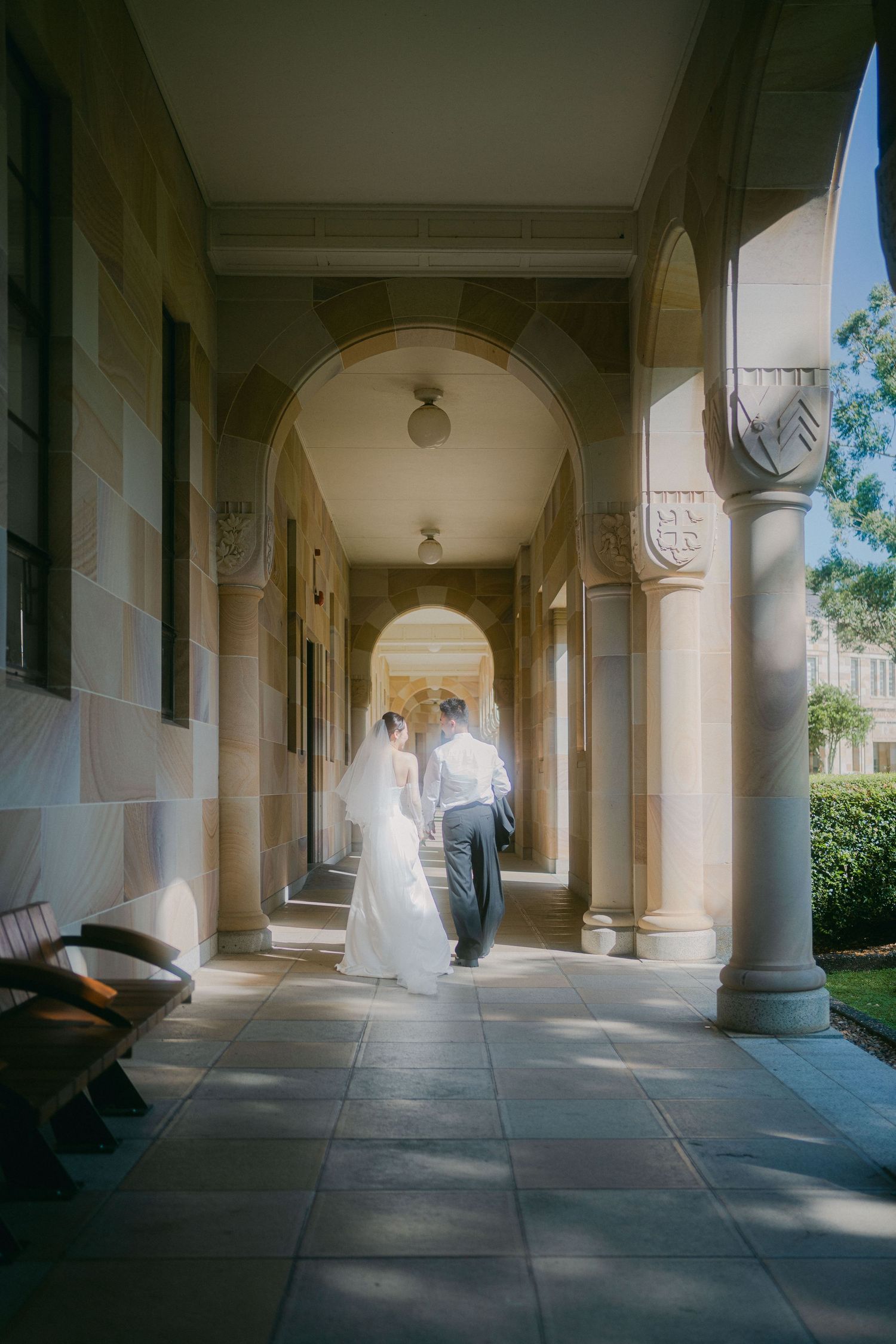 Two figures walk together down an elegant arched colonnade hallway with stone columns and natural lighting filtering in.