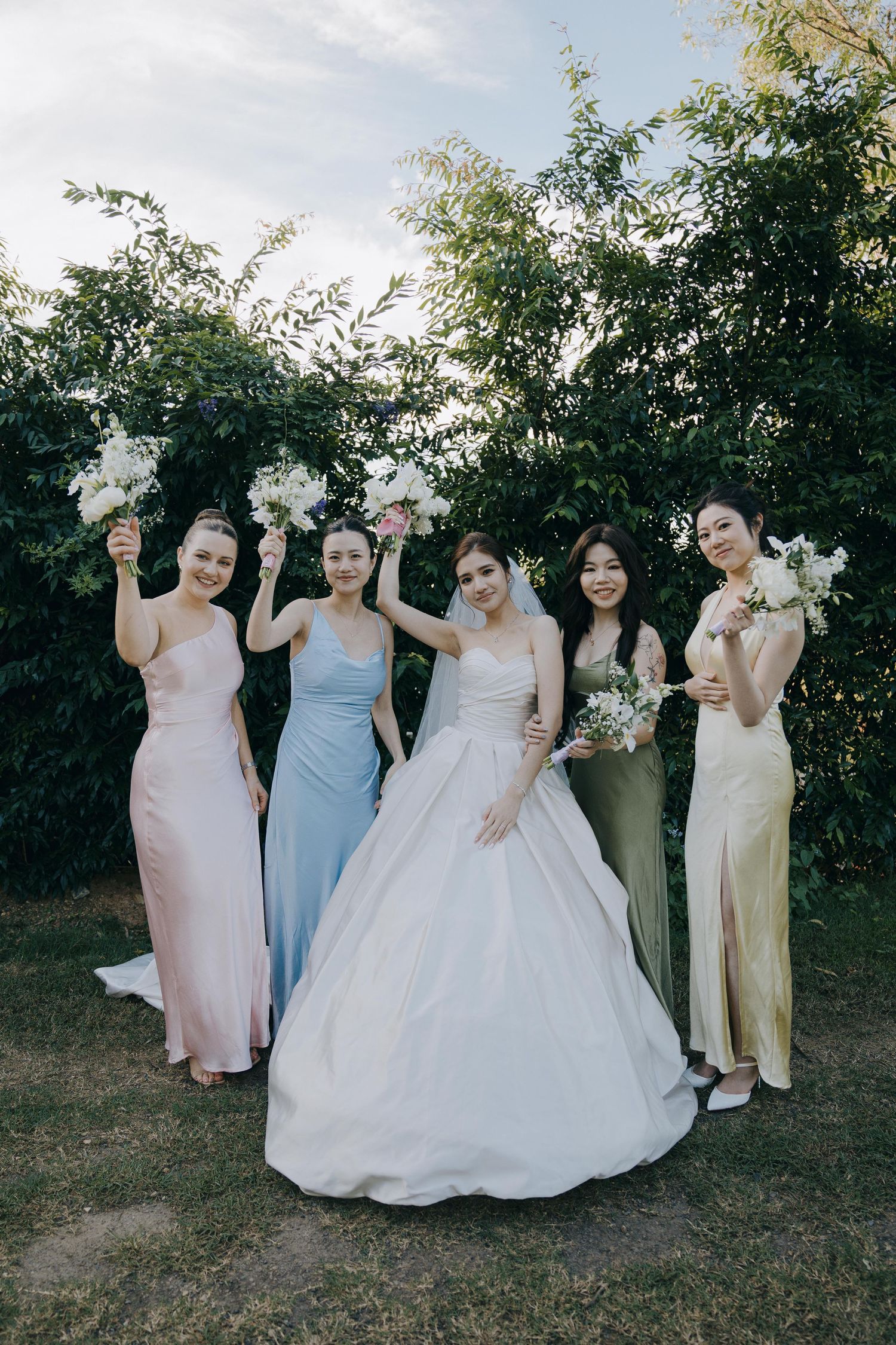 Wedding party in pastel bridesmaids dresses and white bridal gown pose together outdoors with bouquets against greenery.
