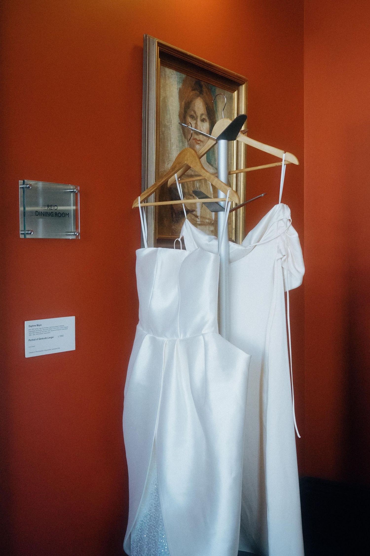 White wedding dresses hanging on hangers against a terracotta-colored wall with a framed artwork above.