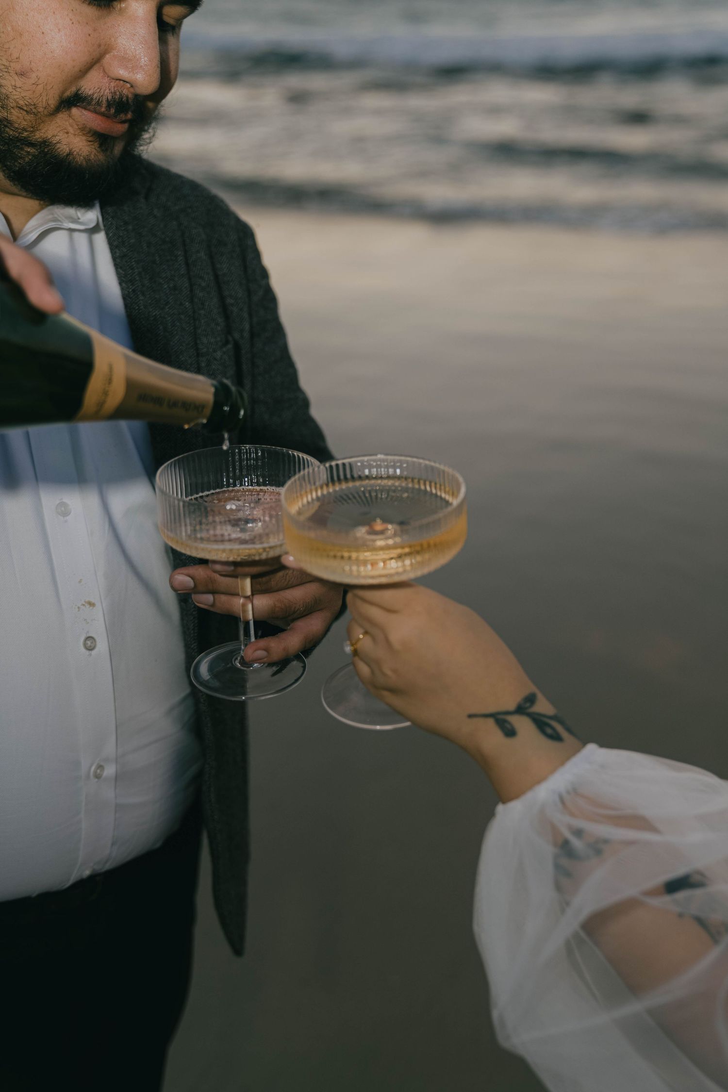 Two people toasting with champagne glasses at sunset on a beach, with one person wearing a dark sweater.