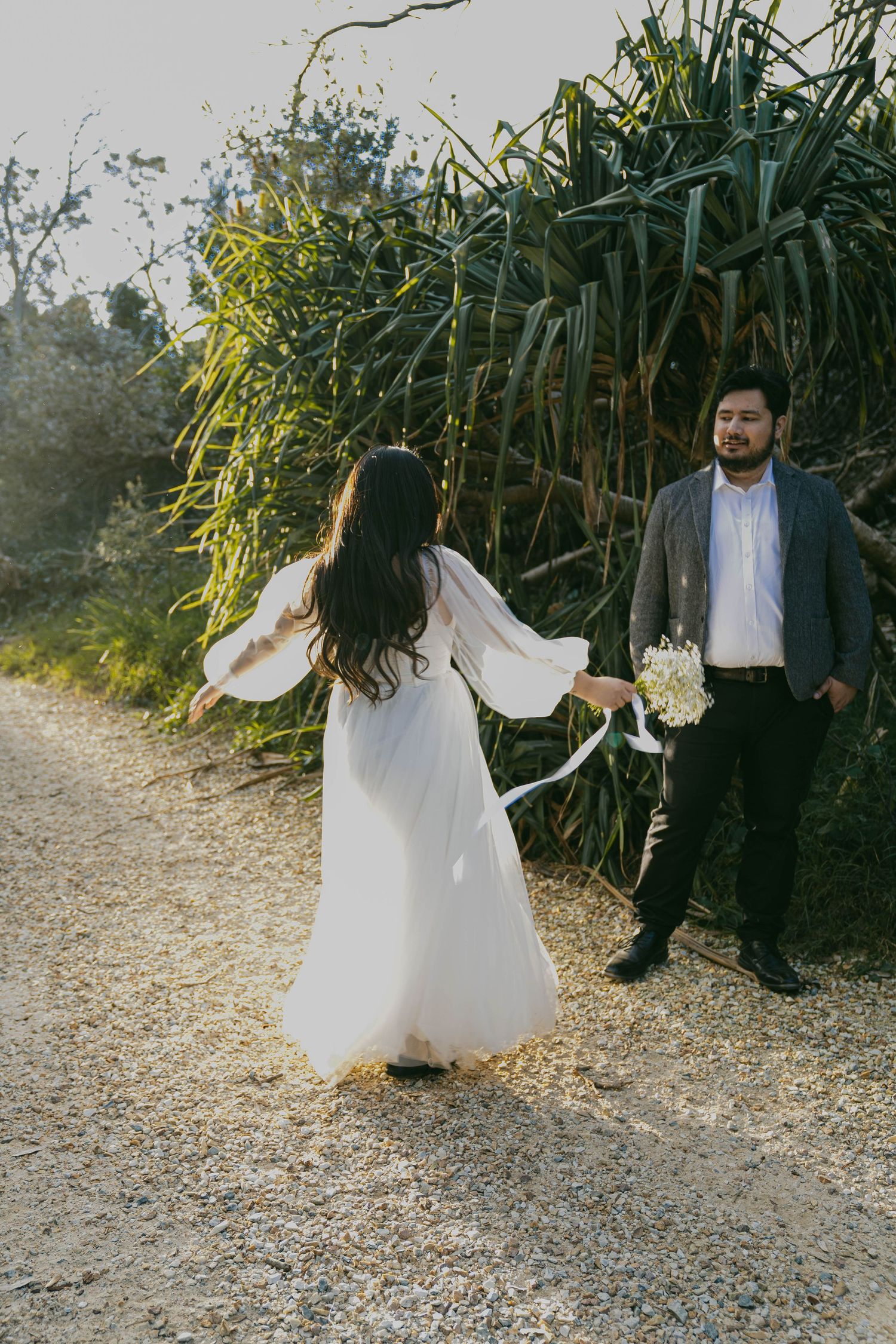 A couple in wedding attire poses on a gravel path with tall bamboo plants in the background during golden hour.