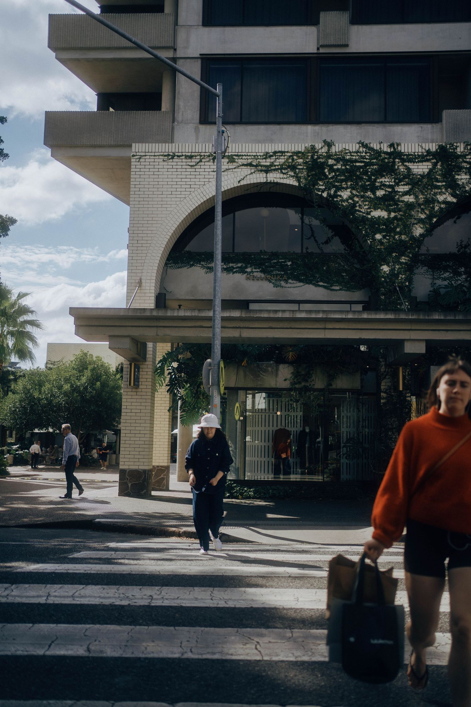 People walk through a modern urban plaza with beige buildings and overhanging balconies on a sunny day.