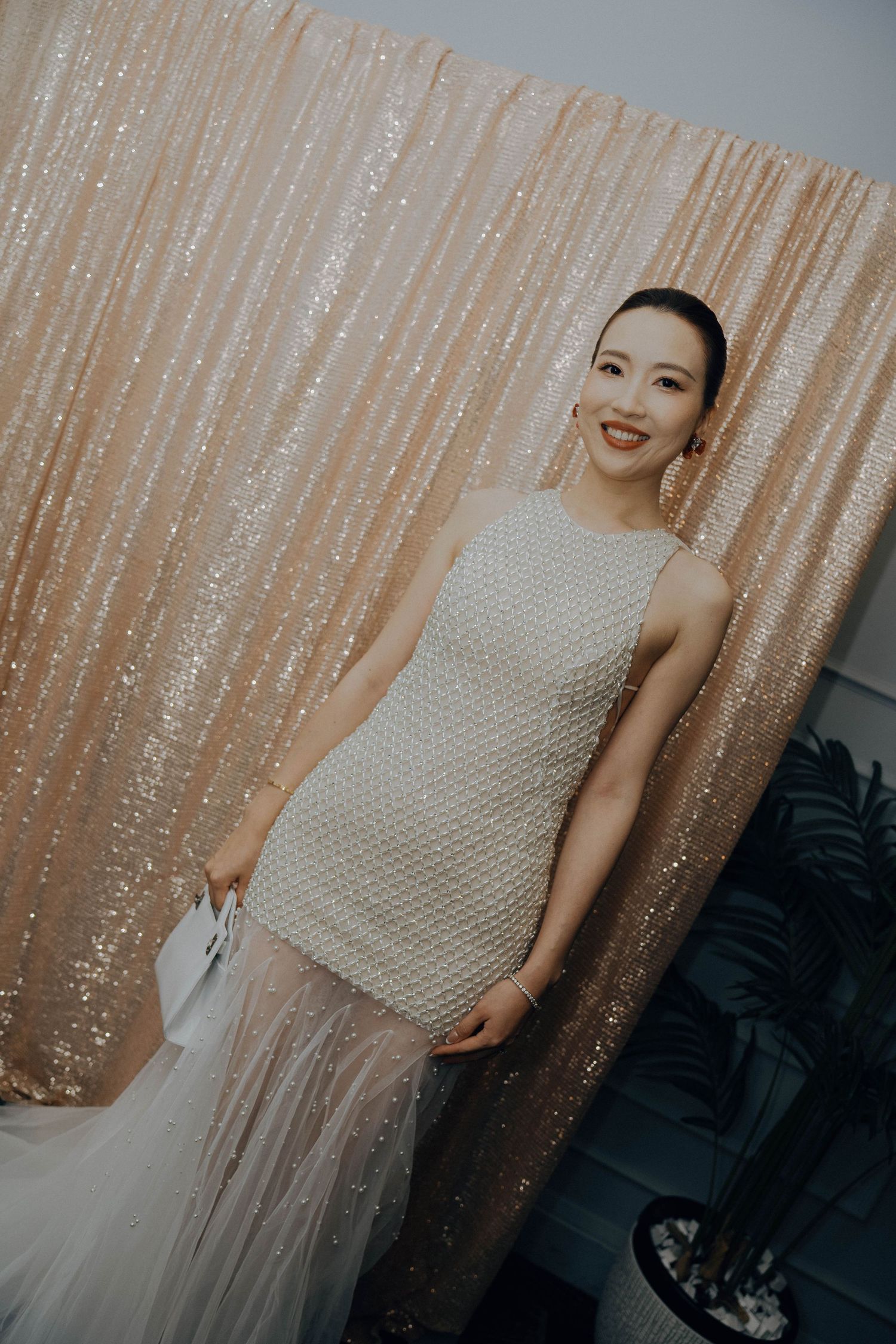 A person in a sleeveless white textured dress poses against a shimmering champagne-colored sequin backdrop.