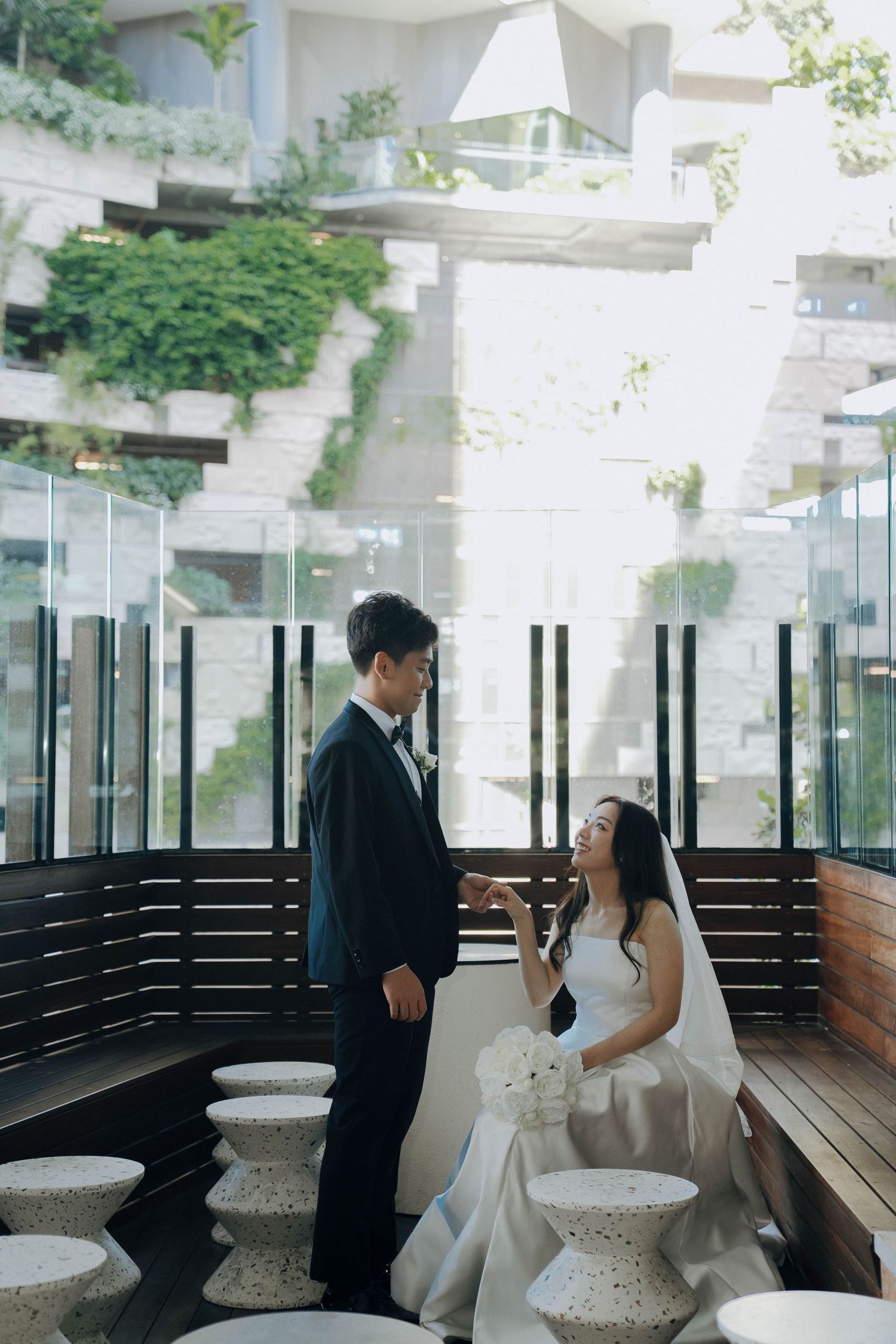 A couple in wedding attire share a moment on a modern terrace with geometric seating and lush greenery.