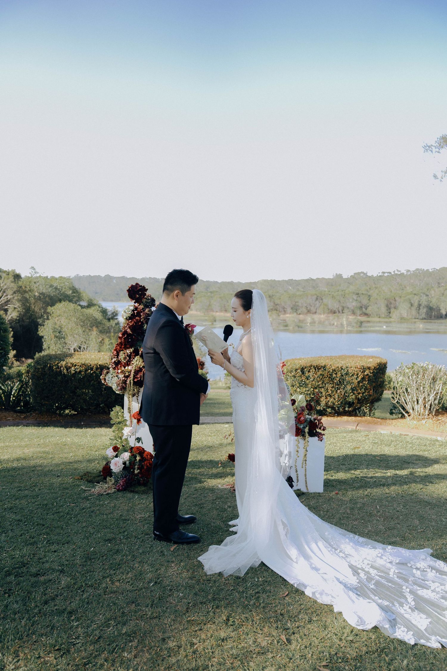 A couple in formal wedding attire shares an intimate moment on a grassy hill overlooking a scenic lake view.