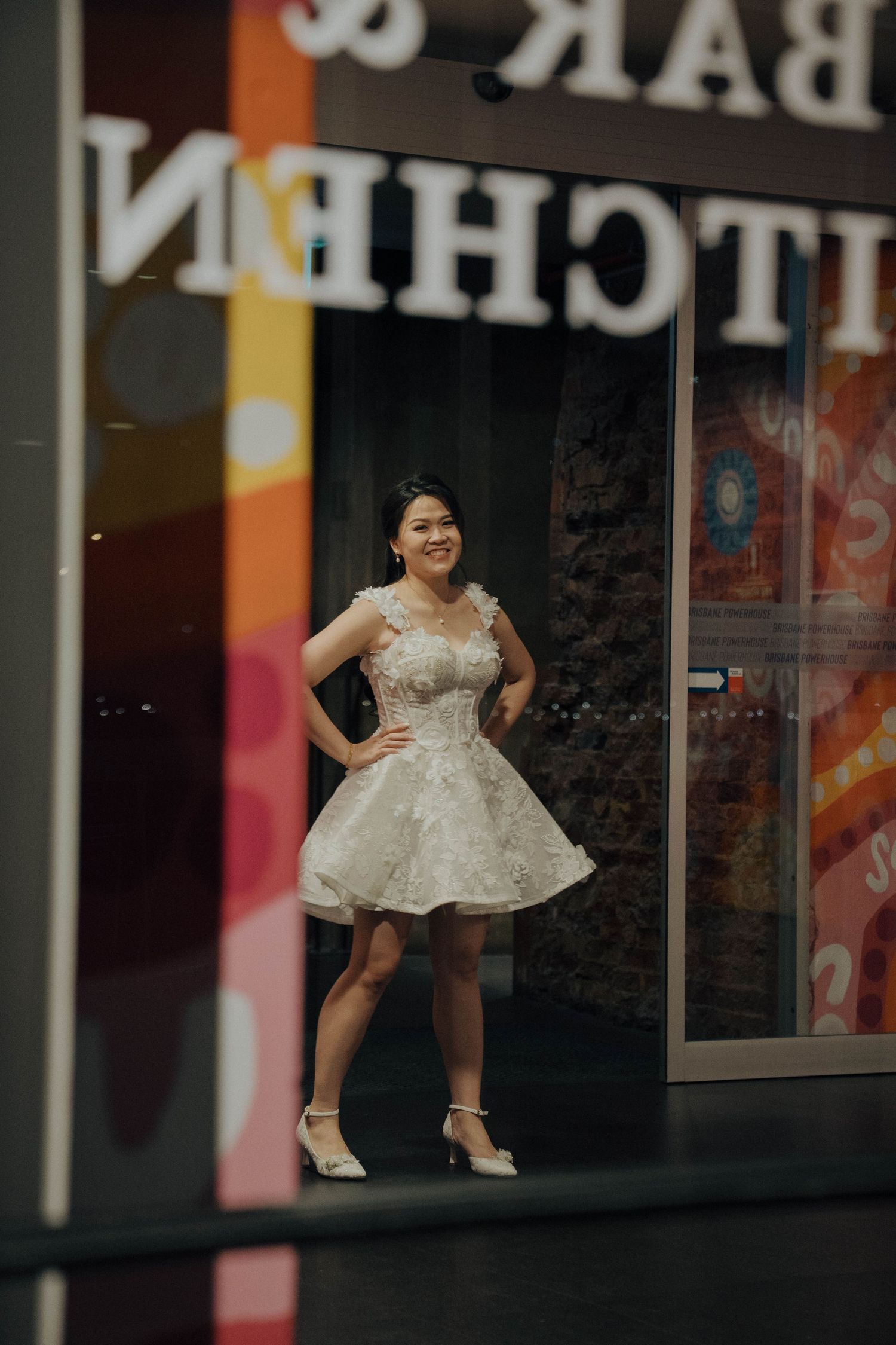 A person in a flowing white dress jumps joyfully in front of a storefront with colorful reflections.