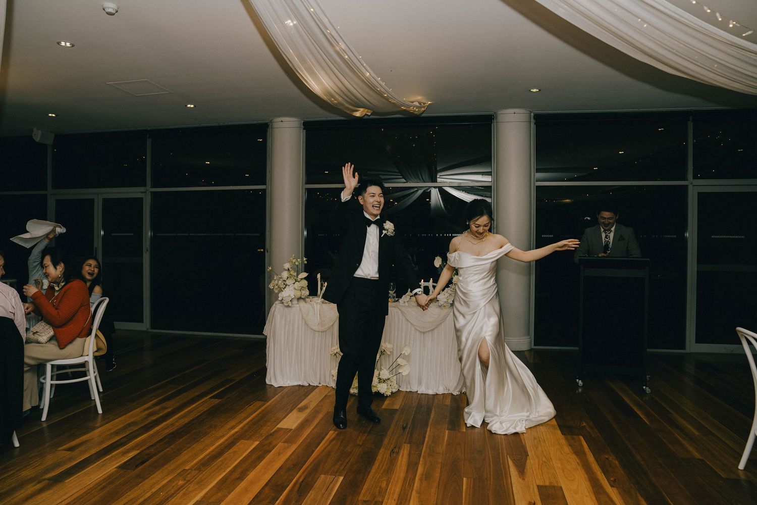 Wedding guests dance and celebrate on a wooden dance floor in an elegant reception venue with draped fabric decor.
