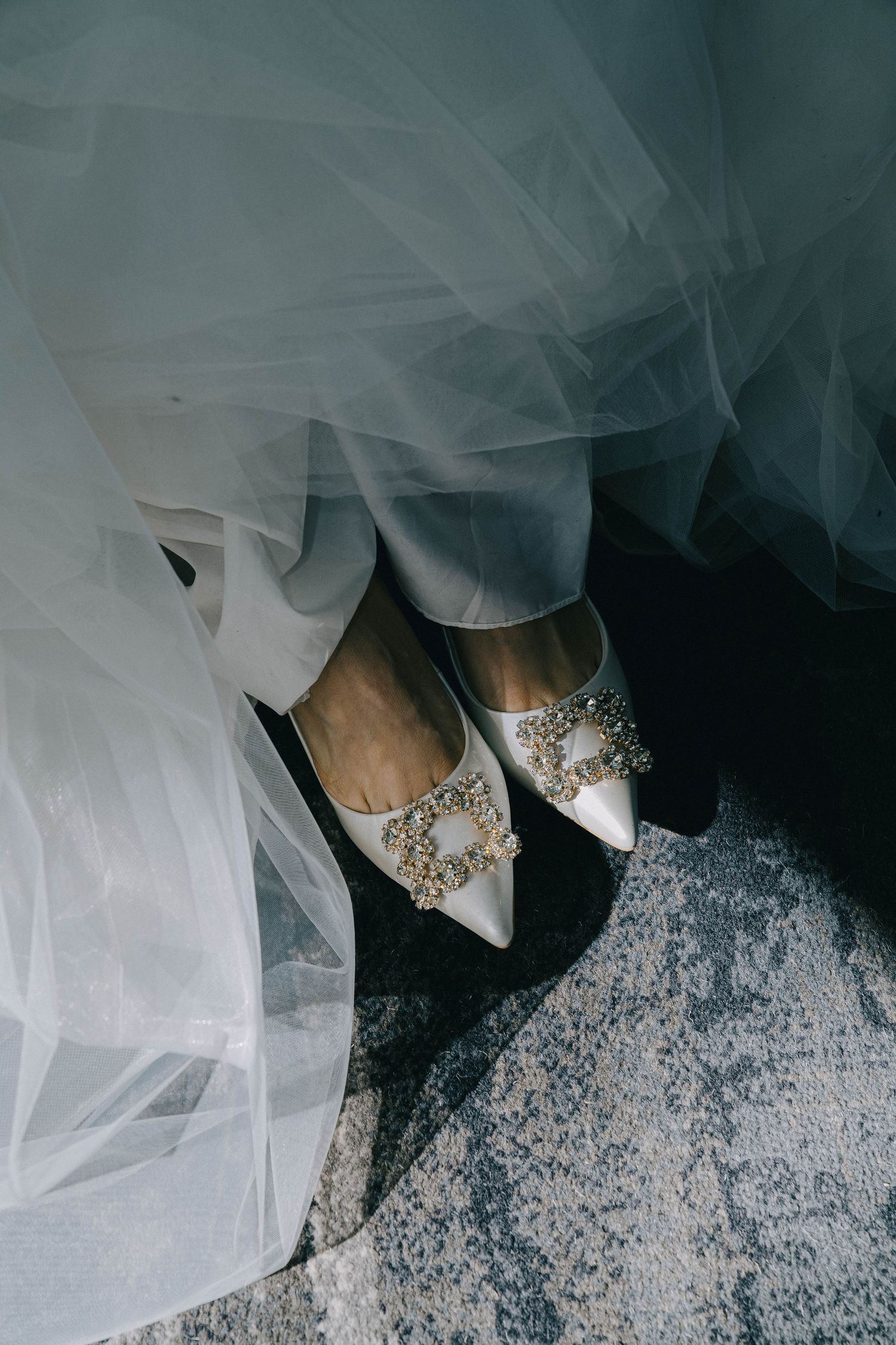 Wedding dress detail showing white tulle fabric and crystal embellished shoes on a textured gray surface.