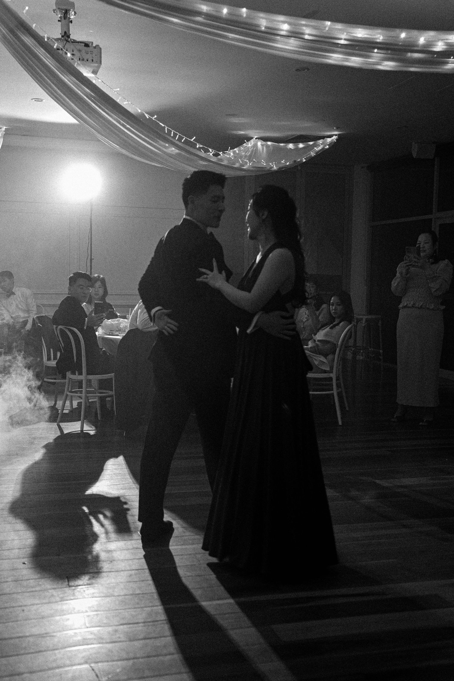 A couple dances together in formal attire at an elegant event with string lights and decorations overhead in black and white.