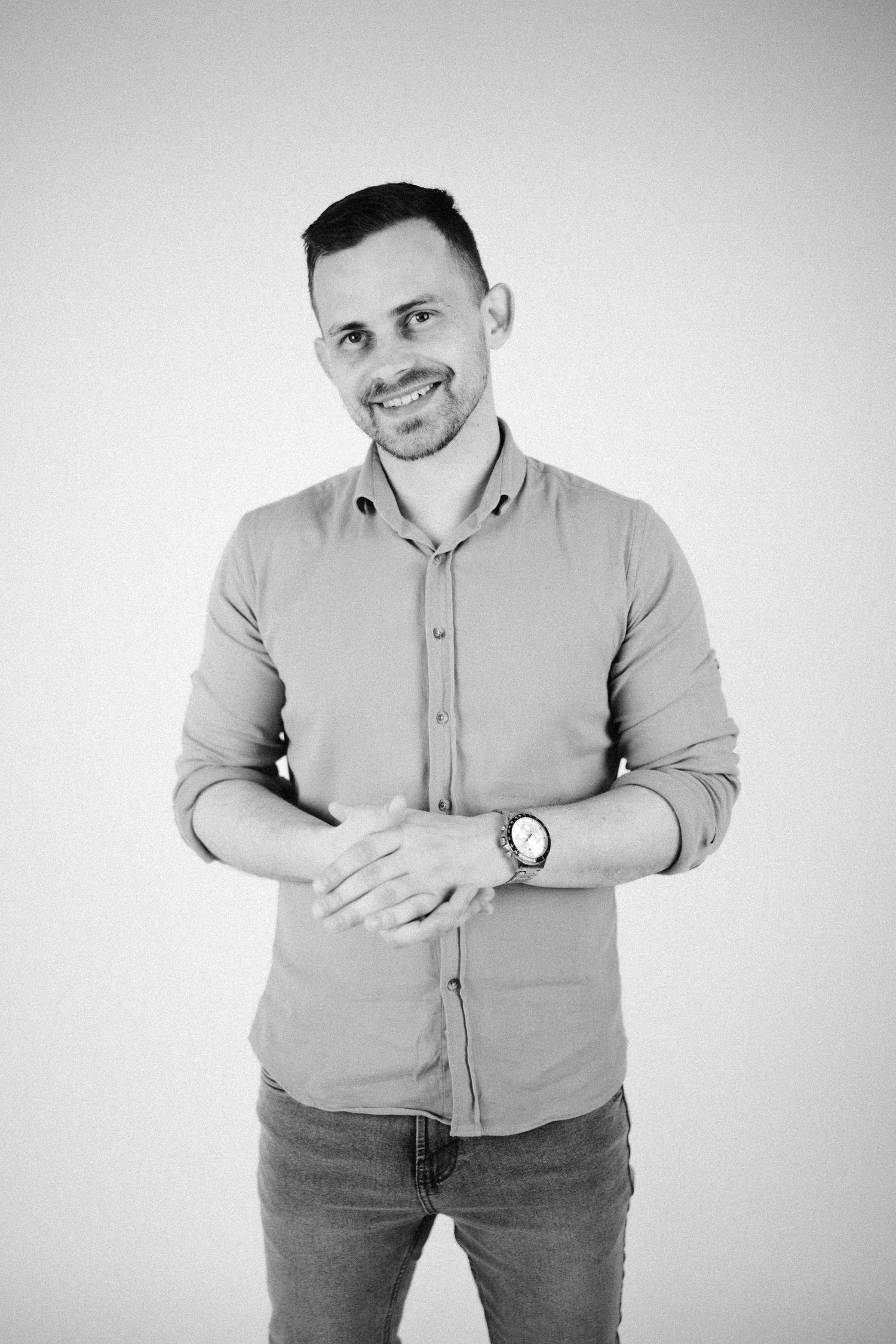 Black and white portrait of someone in a casual button-down shirt and jeans, smiling warmly against a plain background.