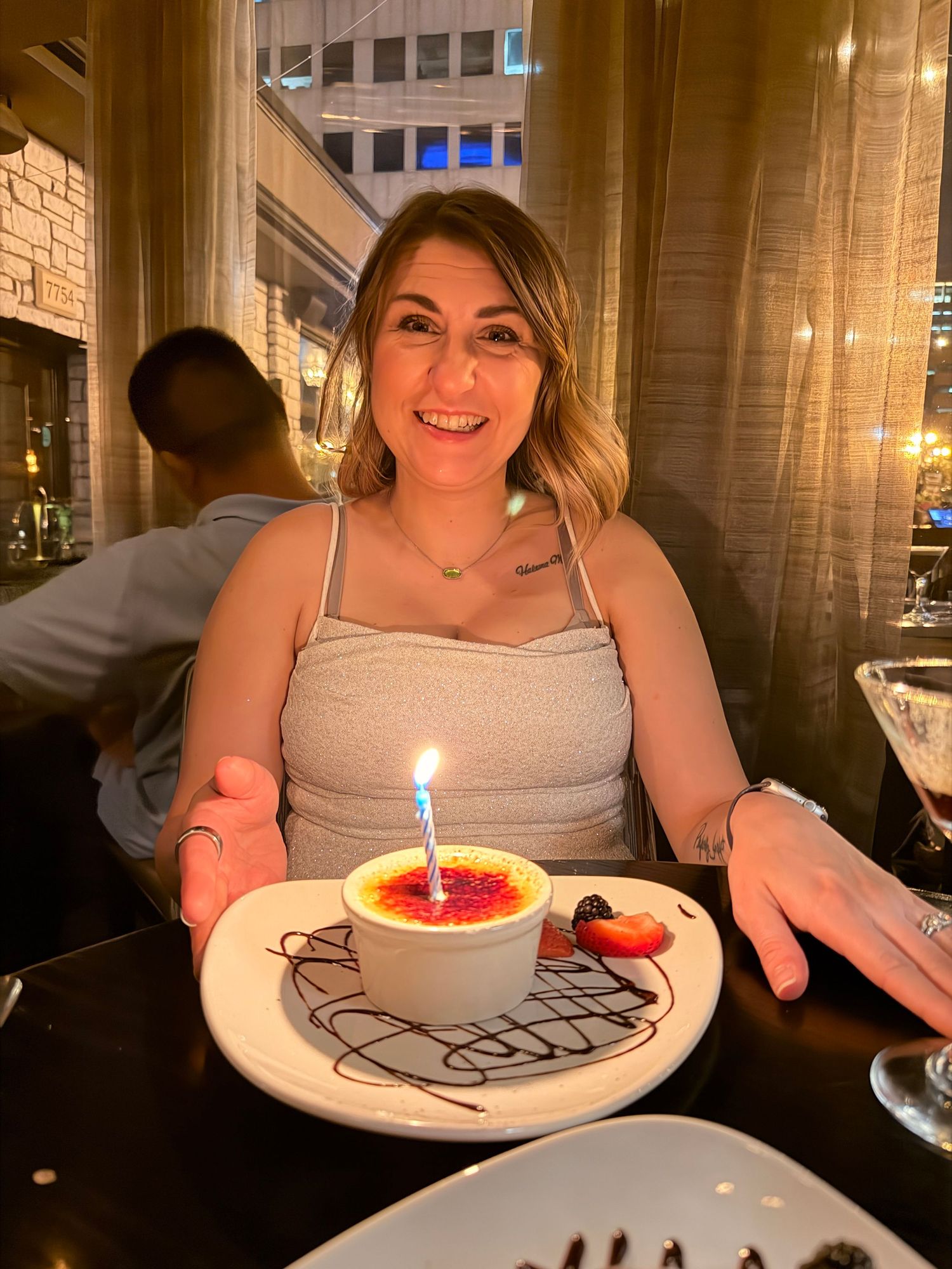 Woman smiling at restaurant table with birthday creme brulee dessert topped with lit candle on white plate.