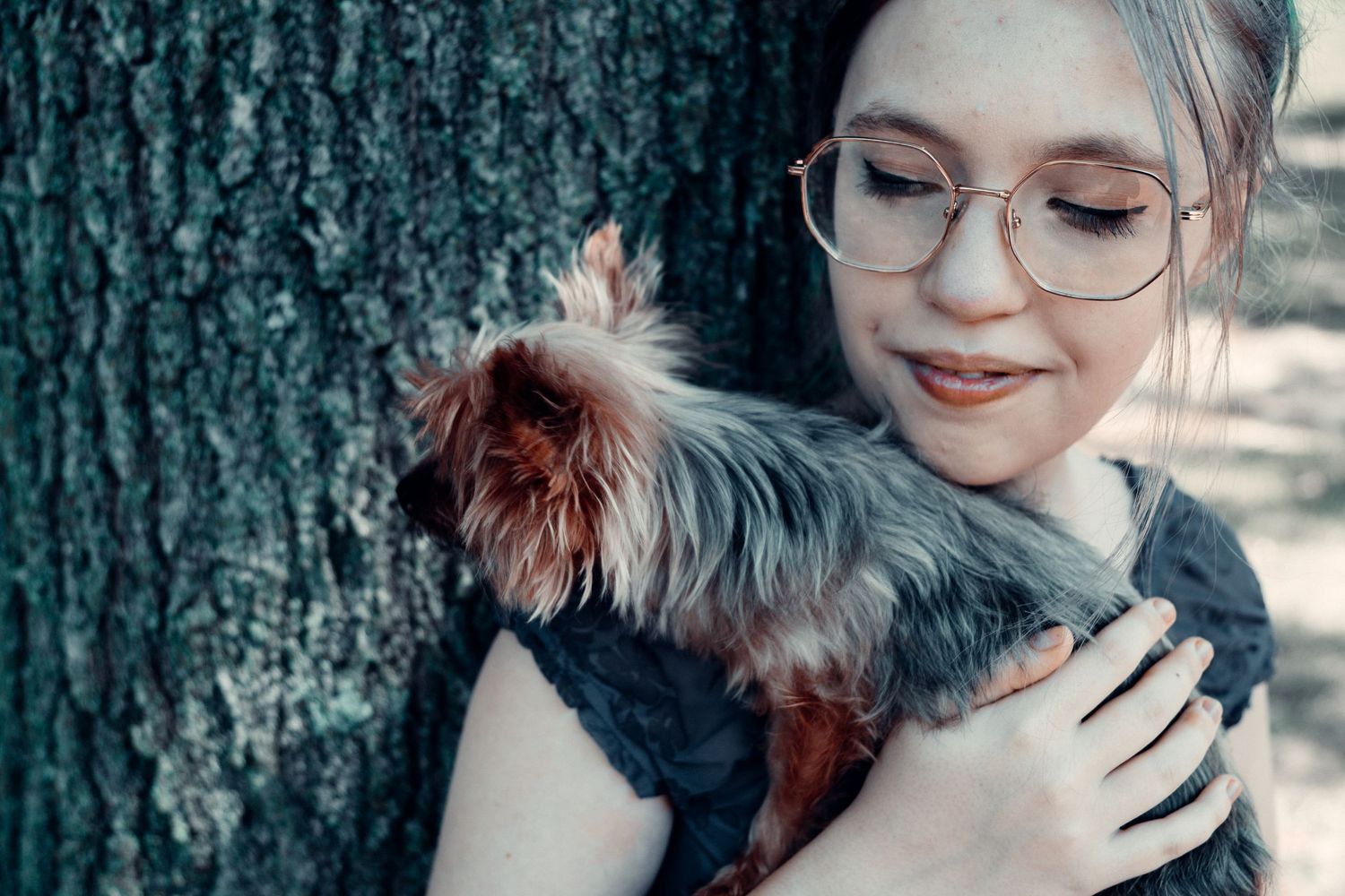 Young woman with glasses hugging a small Yorkshire Terrier dog outdoors near a tree.